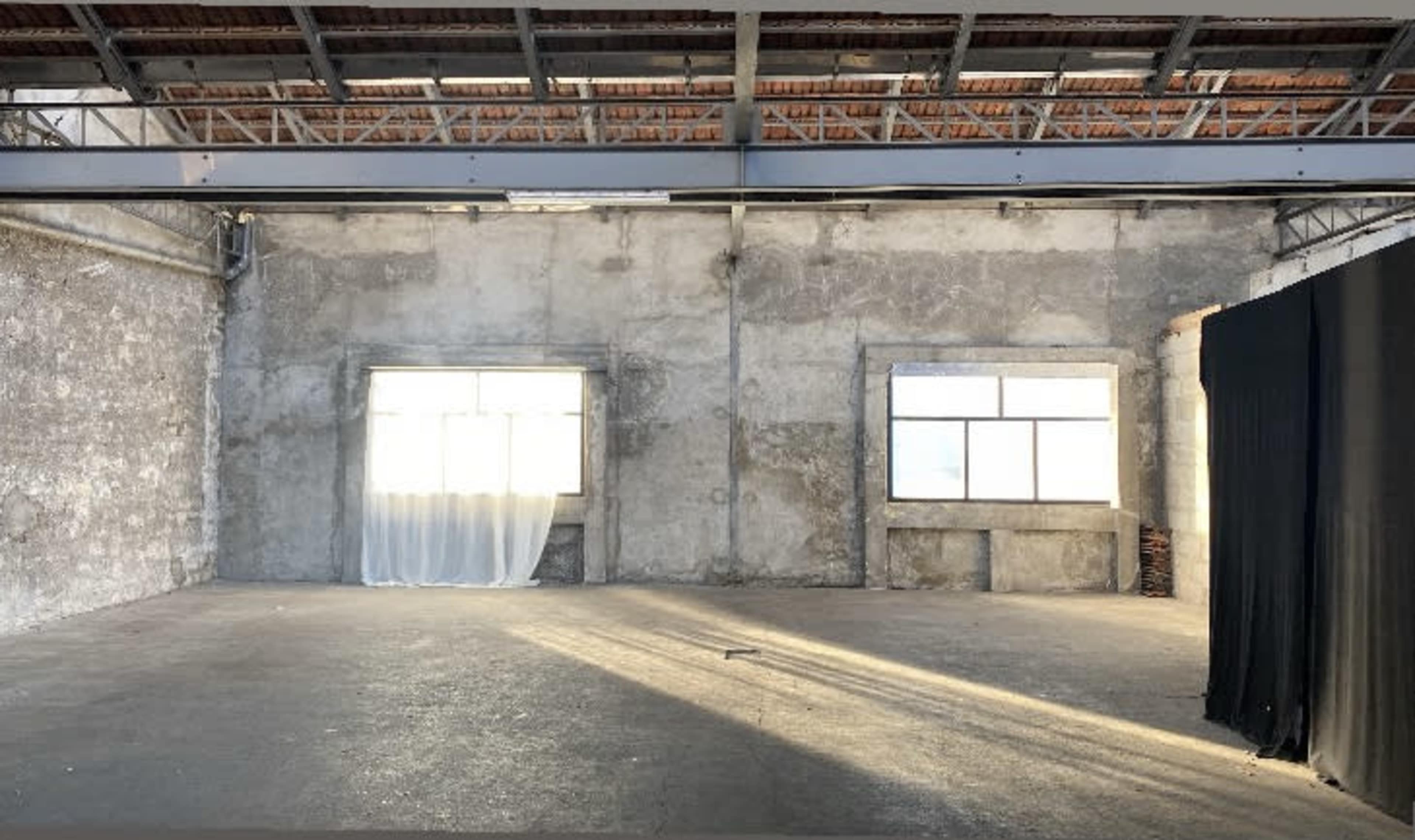 The image shows an empty warehouse interior with bare concrete walls, two large windows, and a section draped with black fabric.