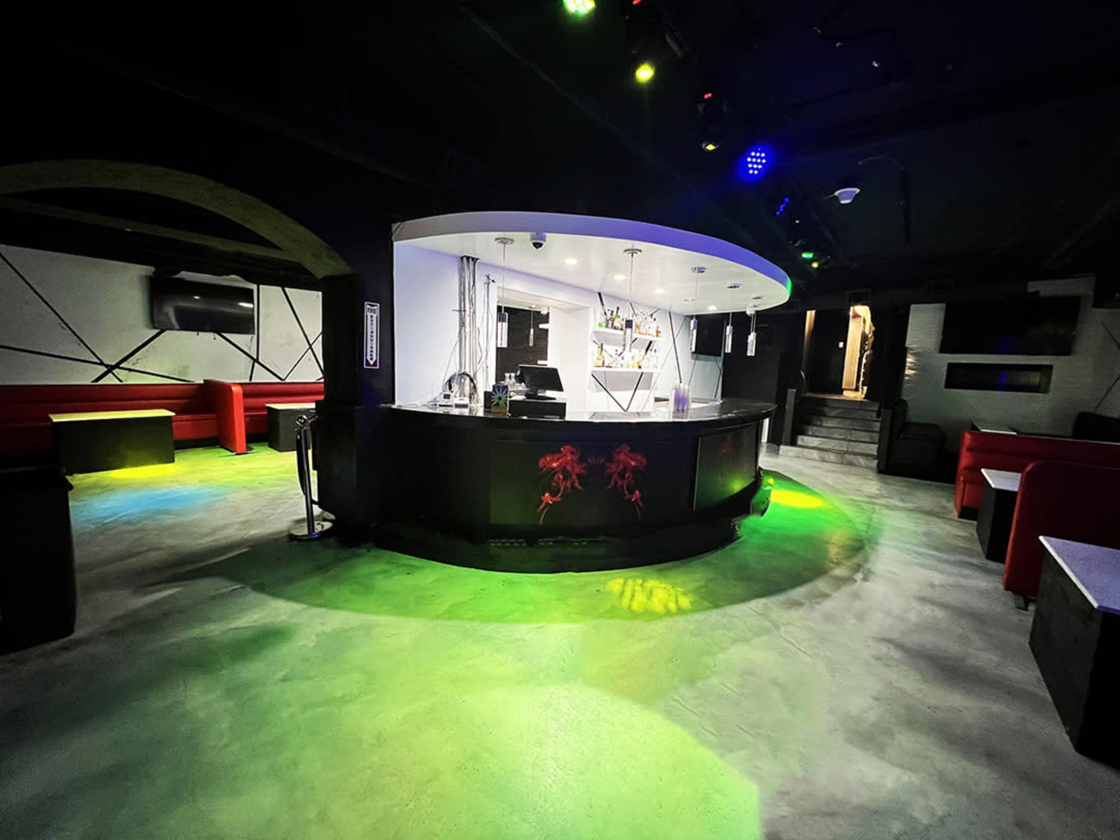 The image shows a dark nightclub interior featuring a circular bar at the center, surrounded by red seating and illuminated by colorful lights.