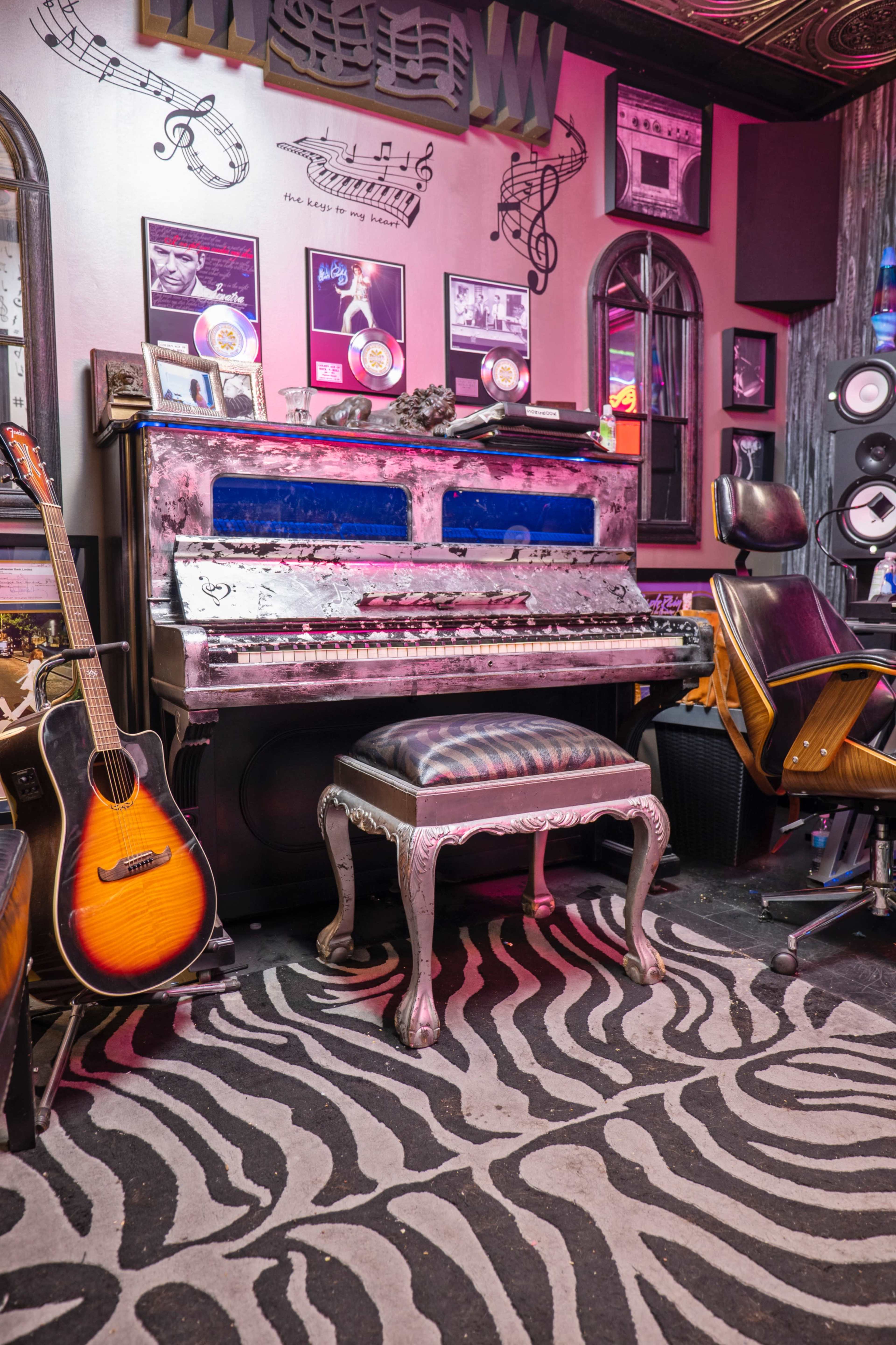 The image features a vintage piano with blue accents, accompanied by an acoustic guitar, set against a wall decorated with musical memorabilia and a zebra-striped rug.