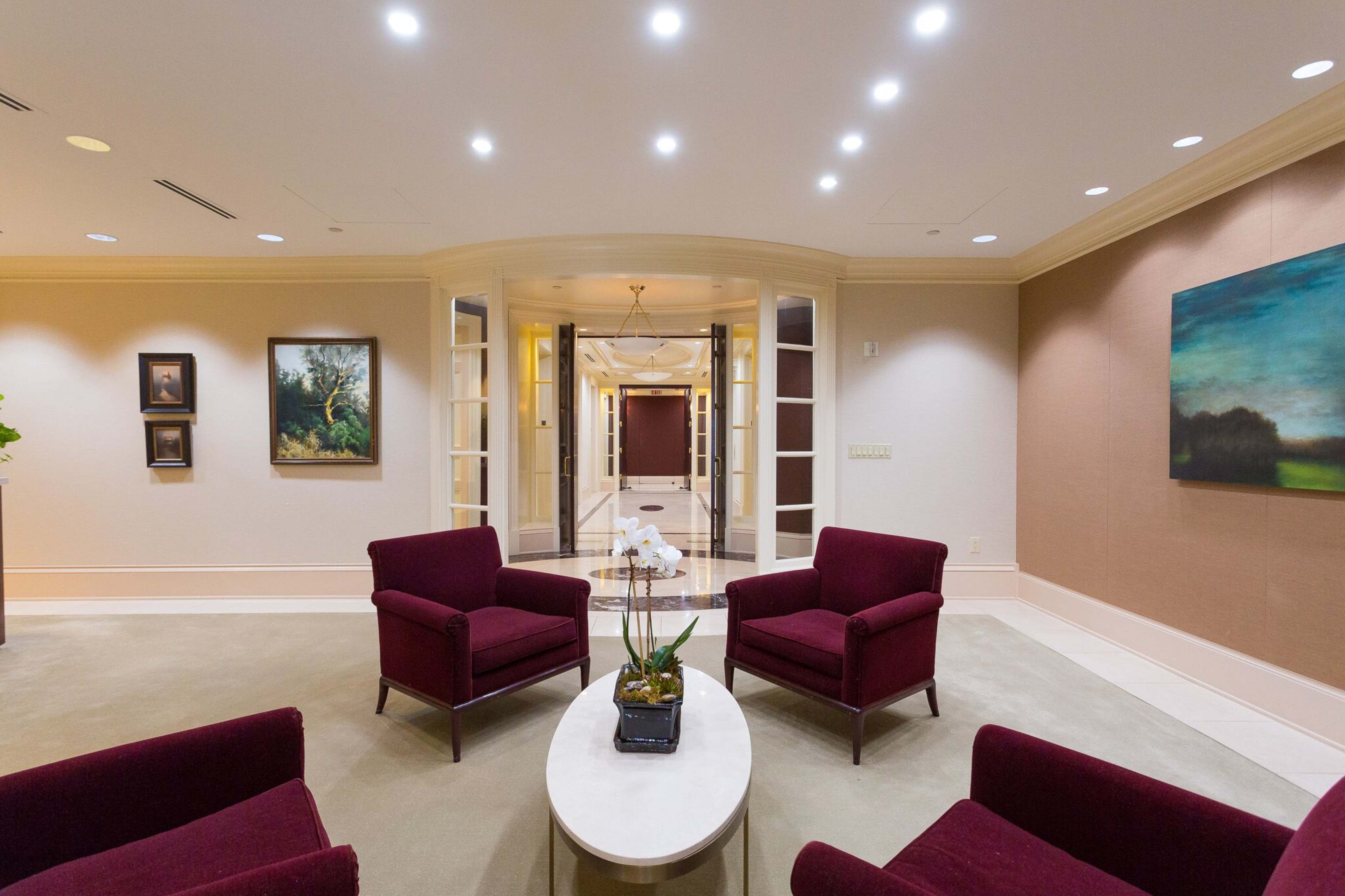 A modern lobby area featuring two maroon armchairs, a coffee table with a plant, and artwork on the walls, leading to an entrance with double doors.