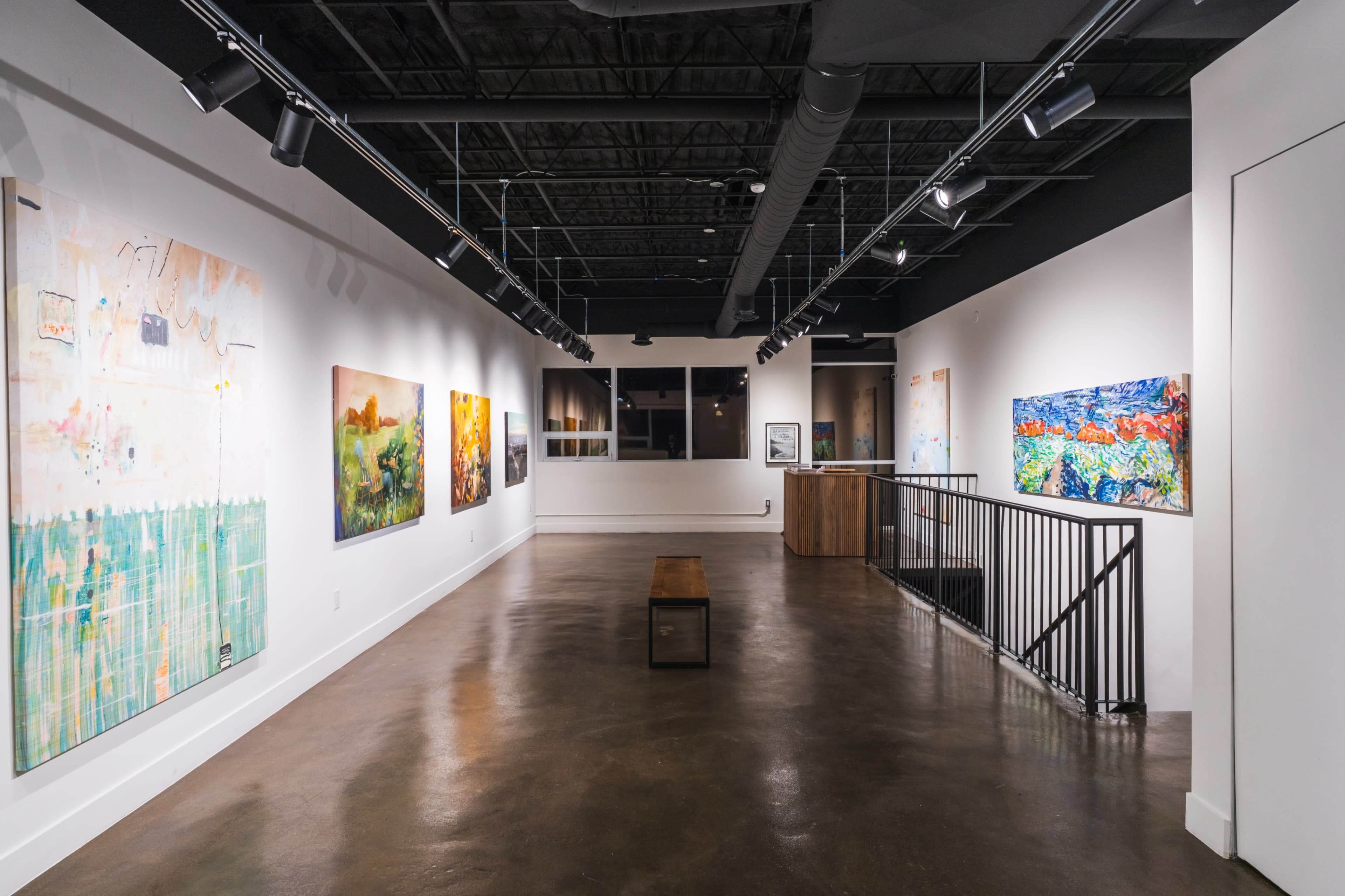 The image shows a contemporary art gallery with several colorful paintings displayed on white walls and a wooden bench in the center of a polished concrete floor.