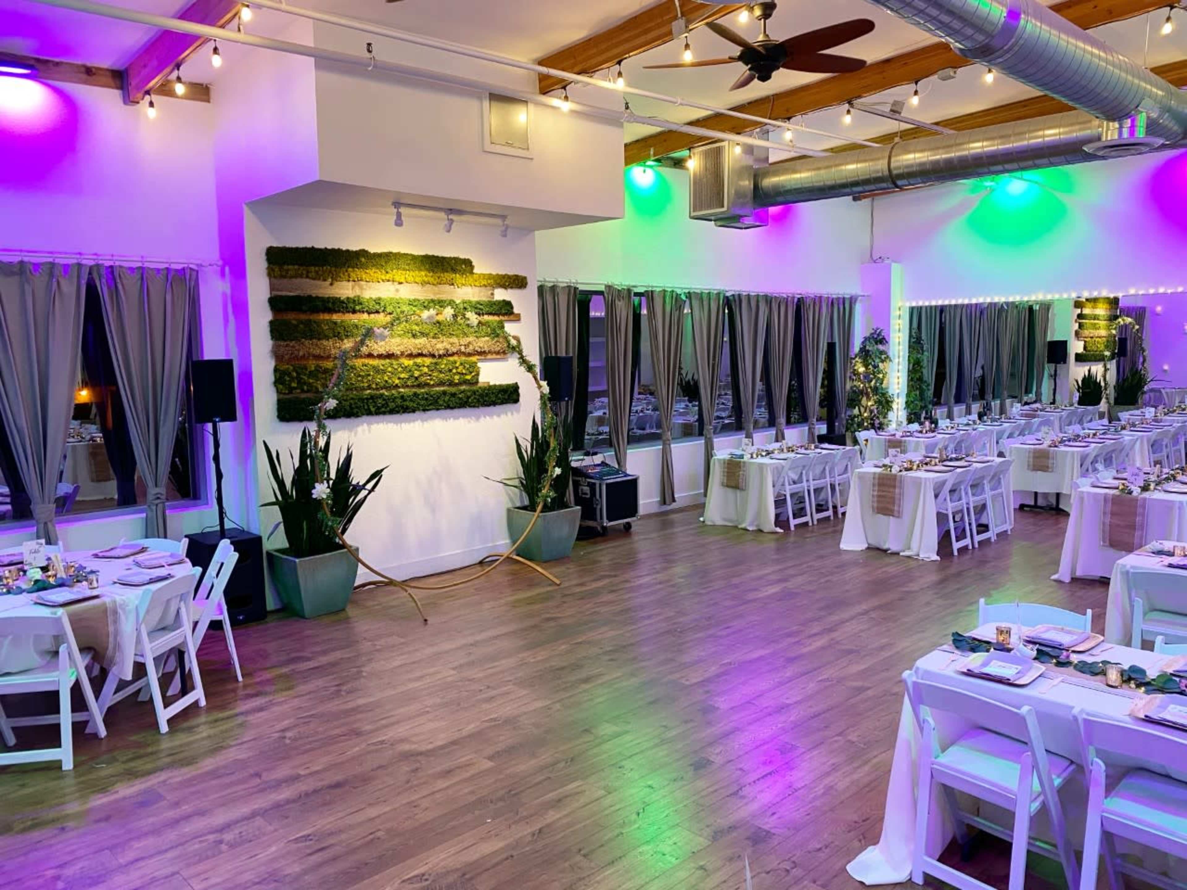 A spacious event room is softly illuminated with green and purple lighting, featuring dining tables arranged neatly with white chairs and a green moss wall as a backdrop.