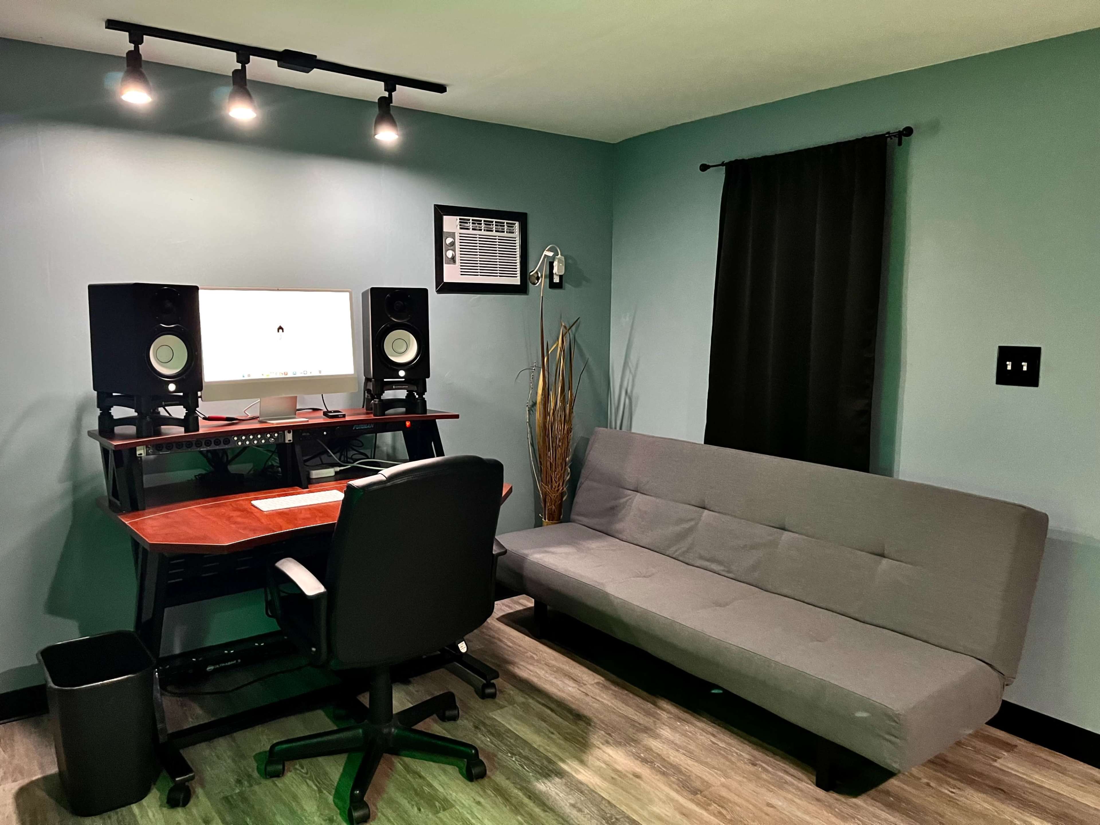 A modern workspace featuring a desk with a computer monitor, speakers, an office chair, and a gray futon against a teal wall.