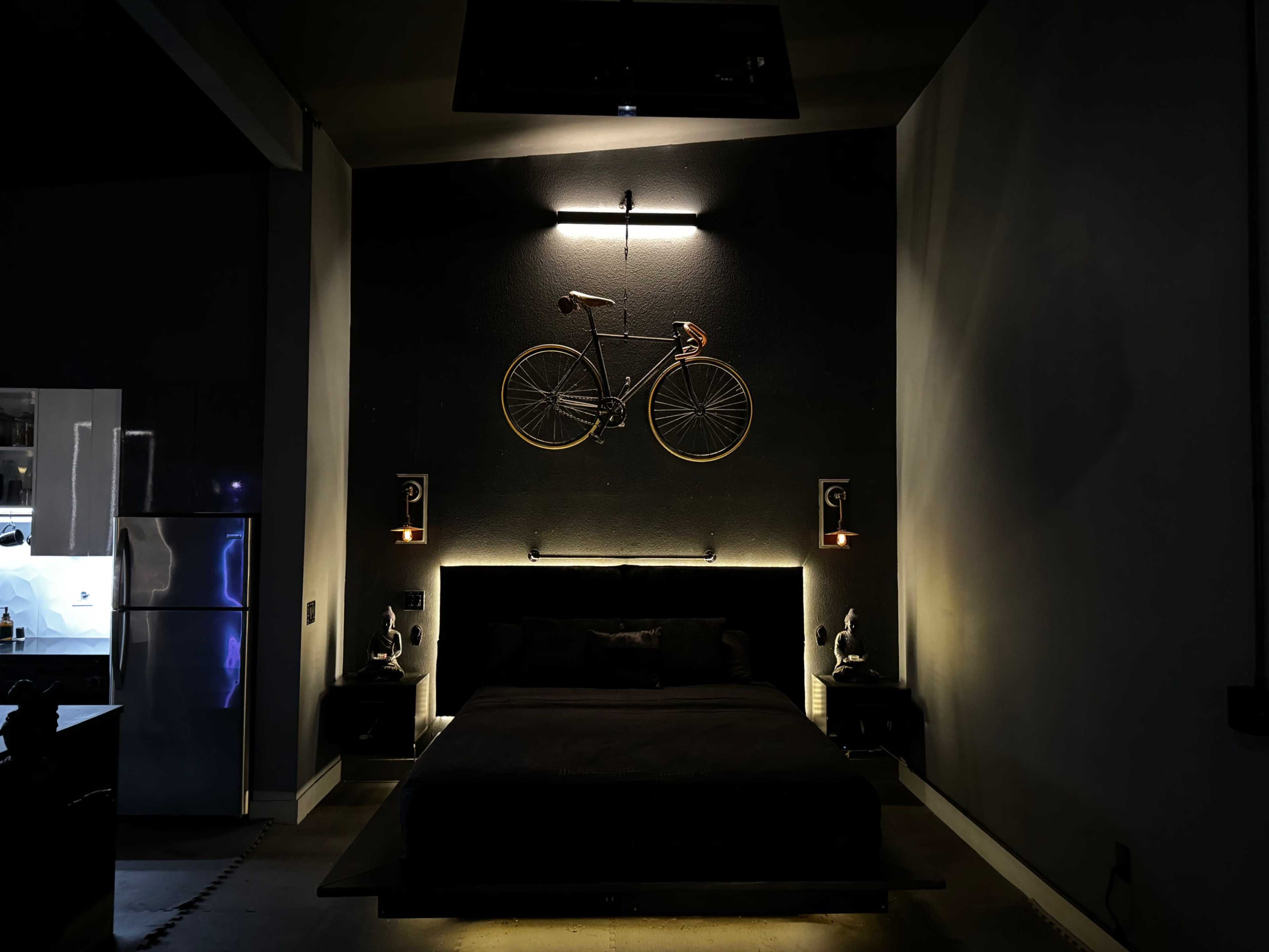 A modern bedroom features a large black bed against a dark wall, with a bicycle mounted above it and minimal lighting from wall sconces.
