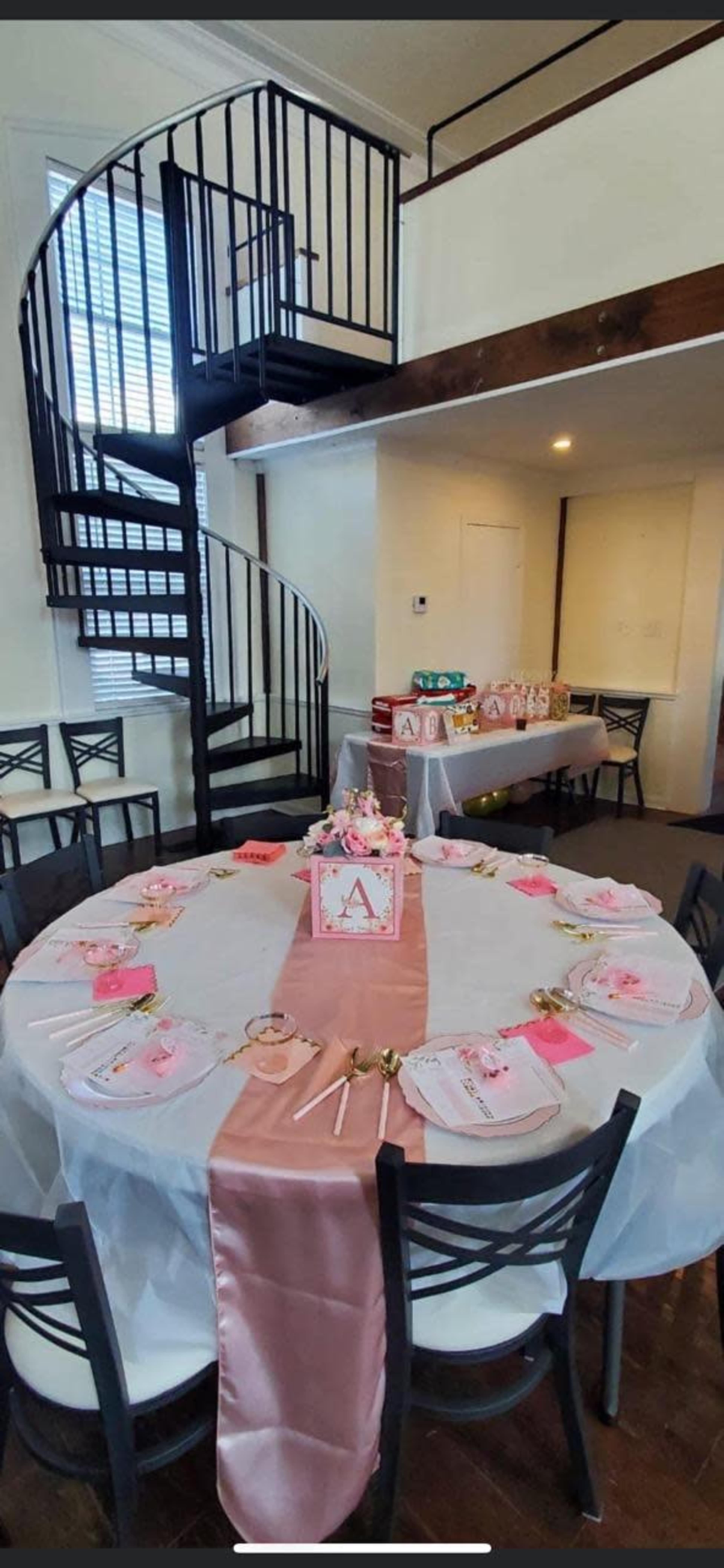 A round table set with white and pink decorations is placed in a room featuring a spiral staircase and a buffet table in the background.