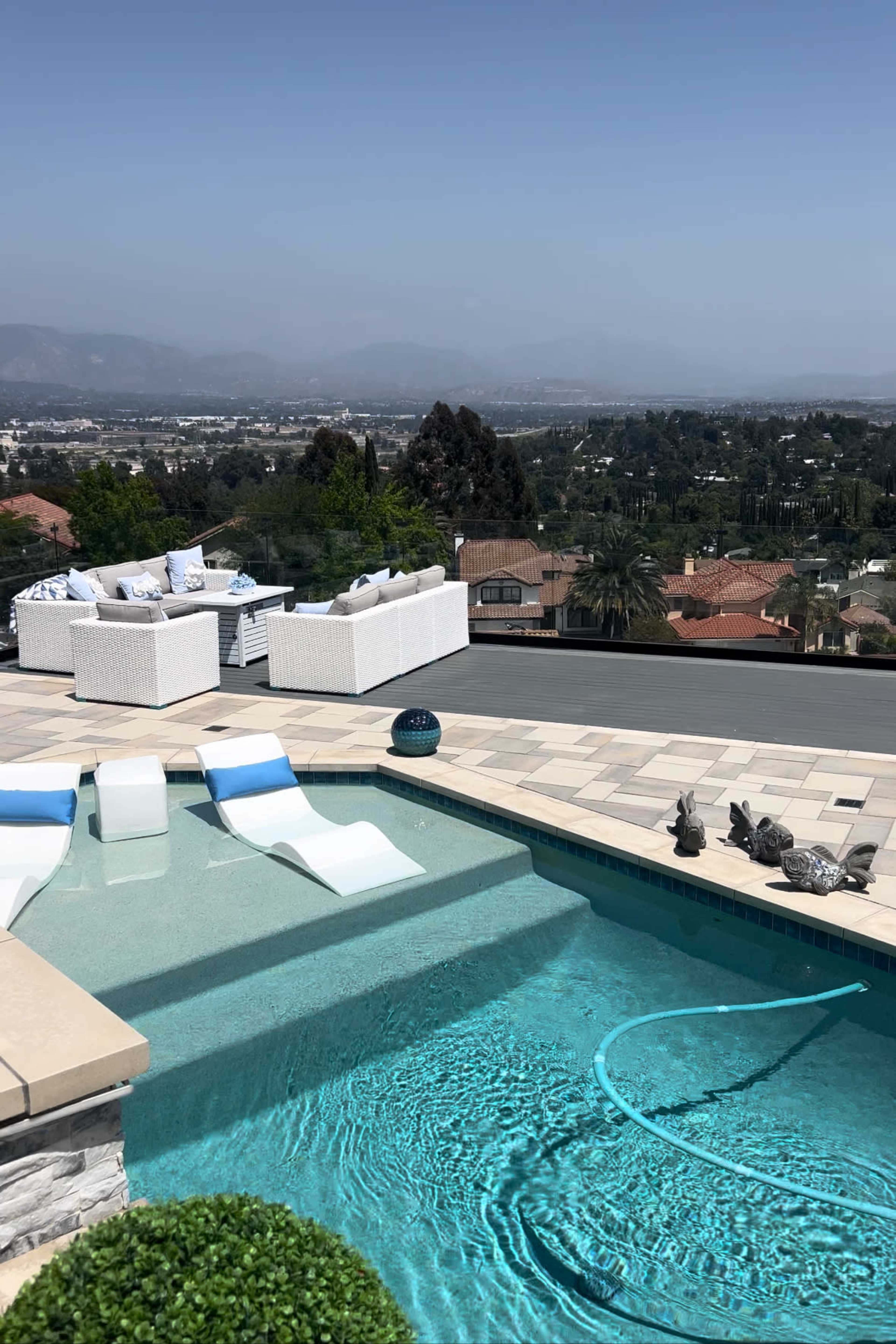 A swimming pool with white lounge chairs is situated on a terrace overlooking a suburban landscape.
