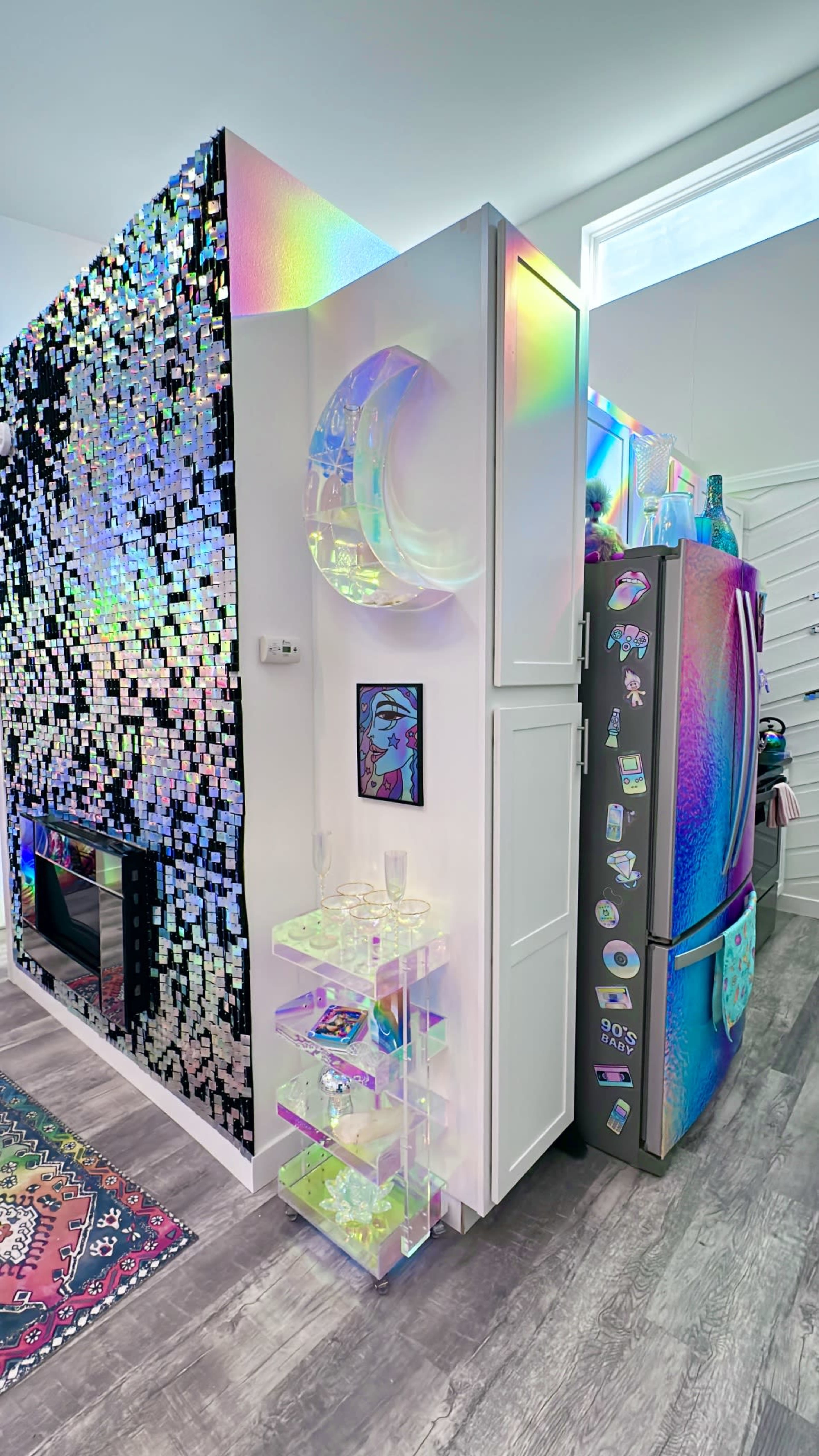 A modern kitchen corner featuring a refrigerator with a reflective, colorful surface, a wall decorated with sequined tiles, and a small transparent shelving unit displaying decorative items.