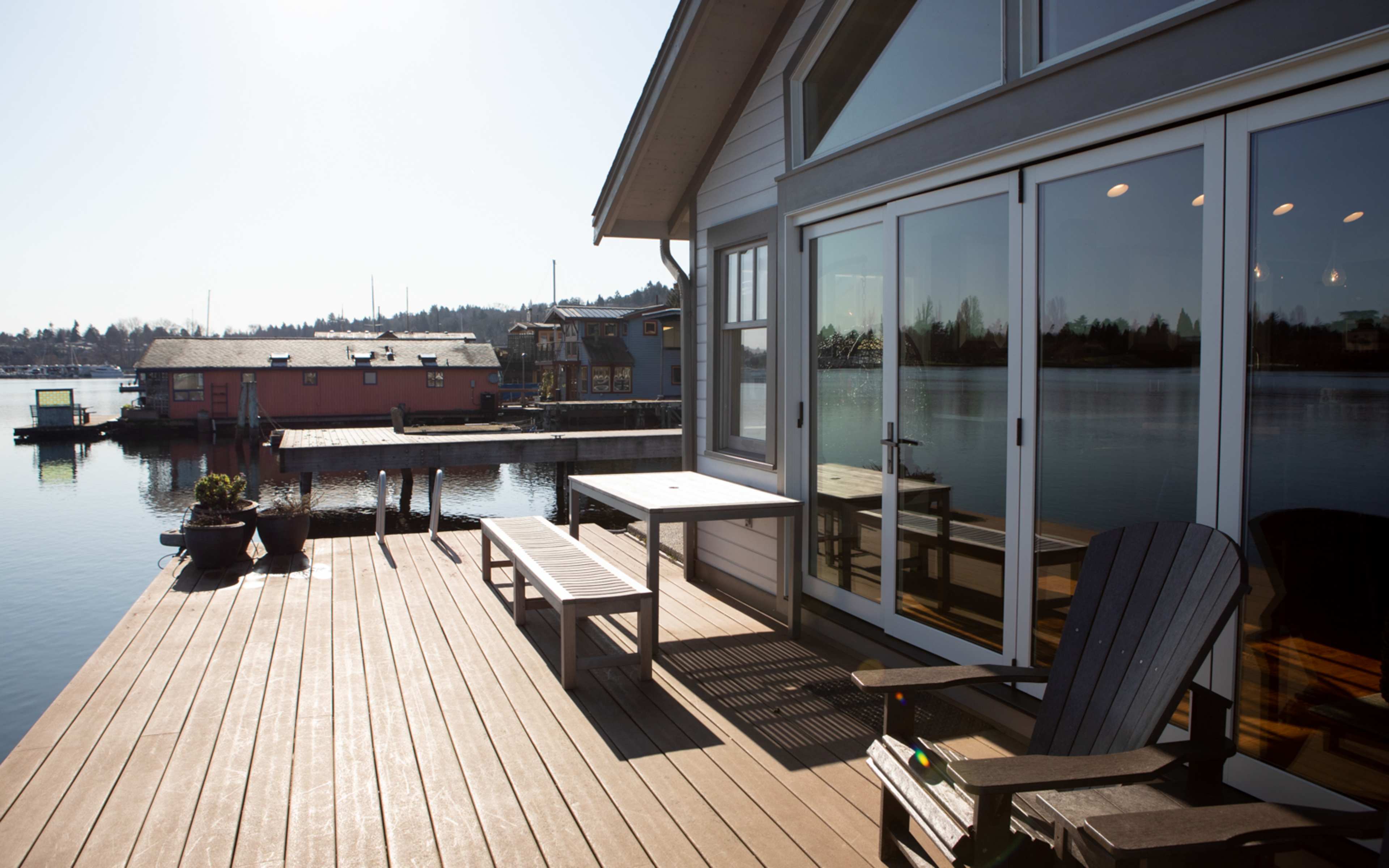 A wooden deck with outdoor furniture overlooking a calm body of water and colorful houseboats in the distance under a clear sky.
