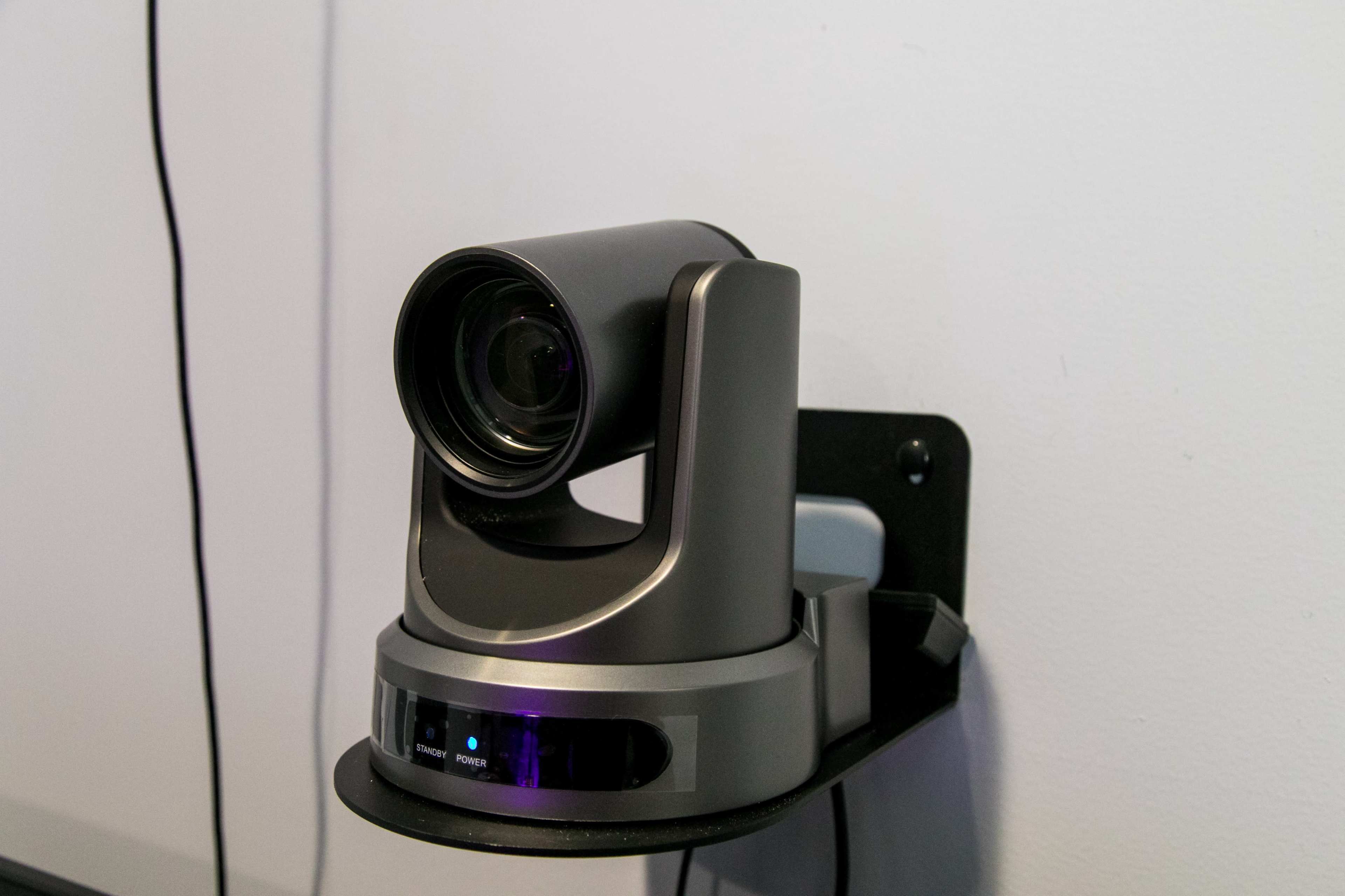A camera is mounted on a wall, featuring a movable lens and a base with a blue indicator light.