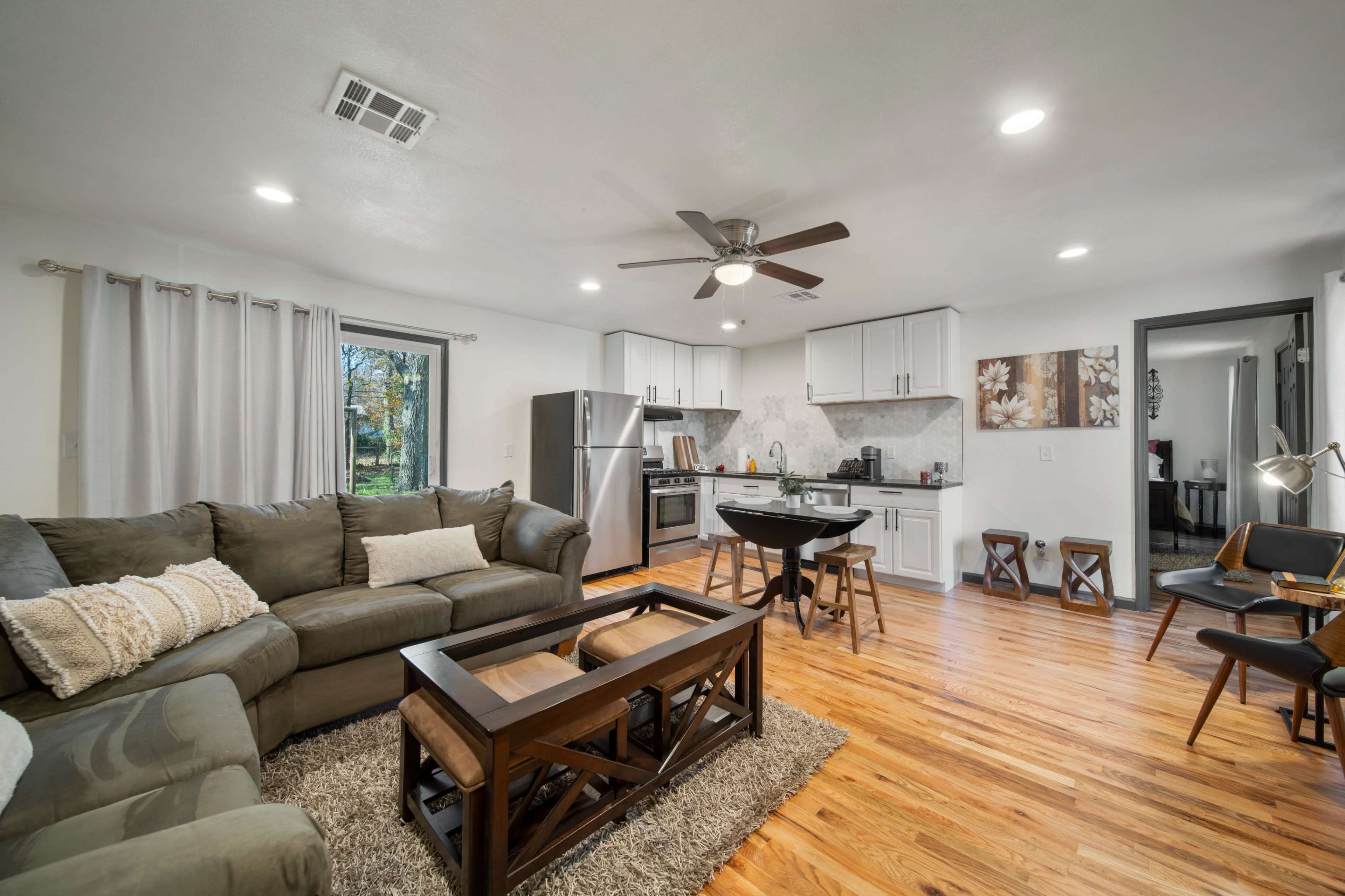 A modern living room features a gray sectional sofa, a wooden coffee table, and a dining area with a round table, surrounded by large windows and wooden flooring.