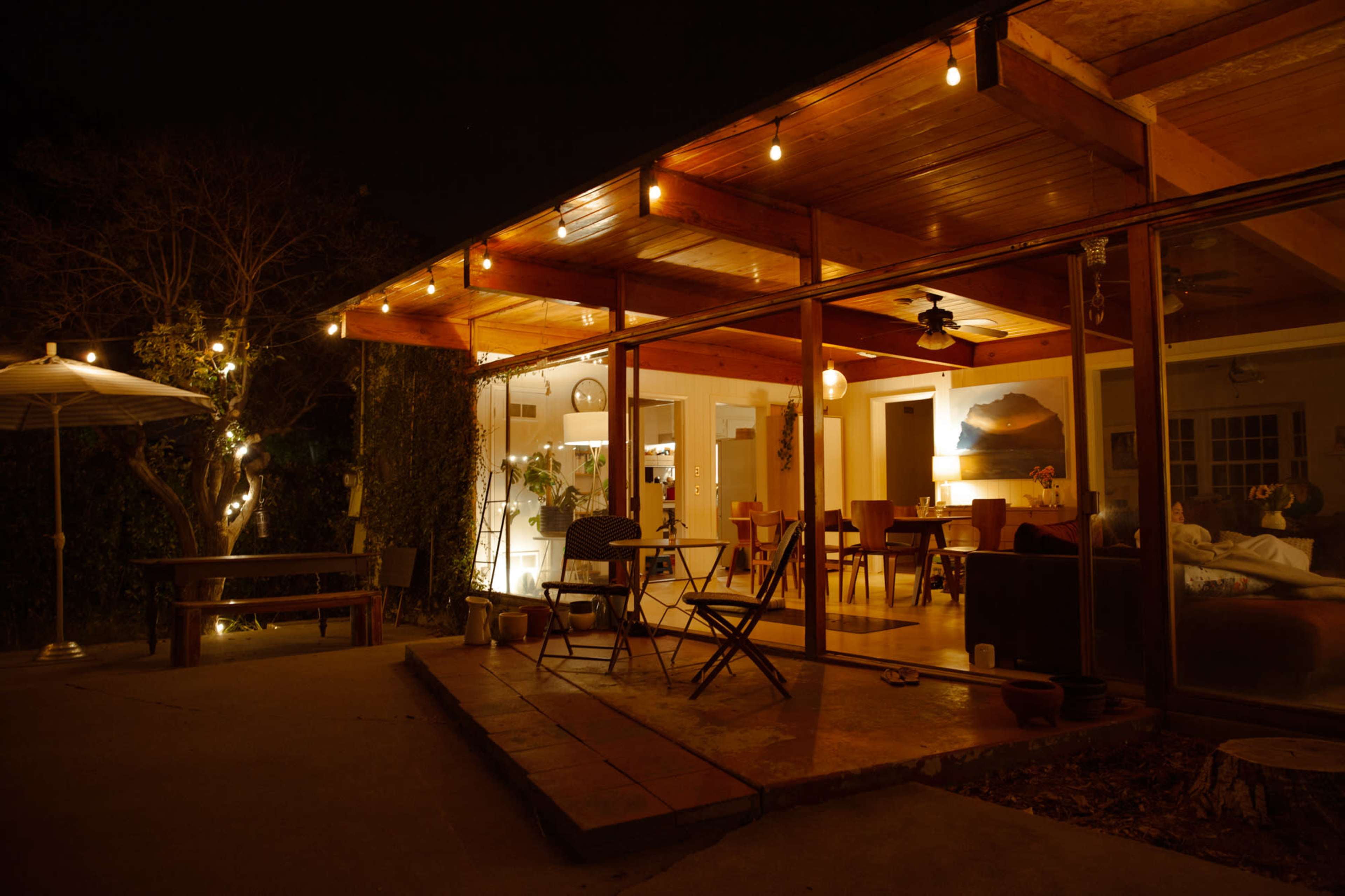 A cozy outdoor patio area features string lights illuminating a glass-enclosed living space with wooden beams and a table set for dining.