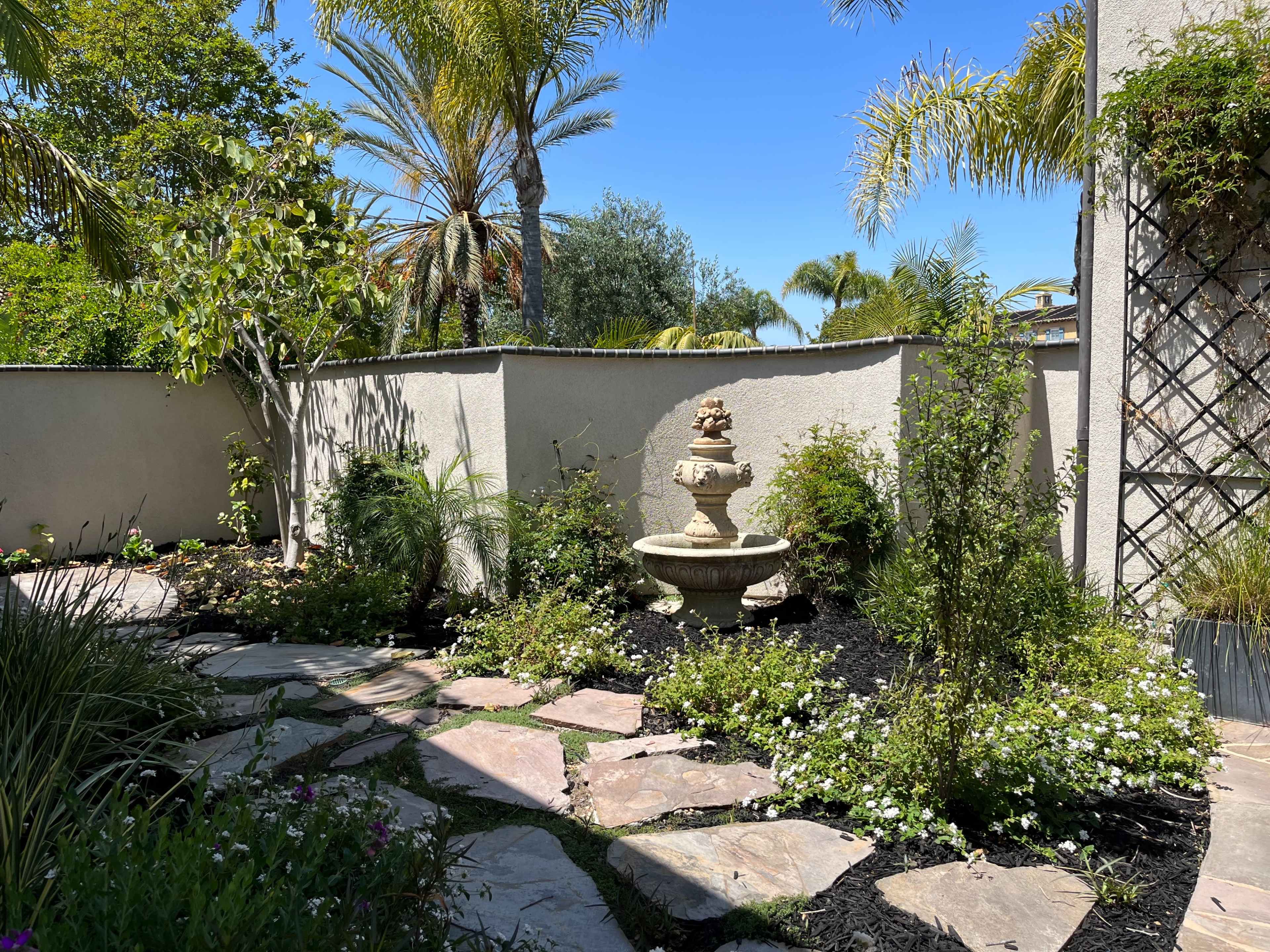 A stone fountain is surrounded by a path of flagstones and lush greenery in a sunlit garden.