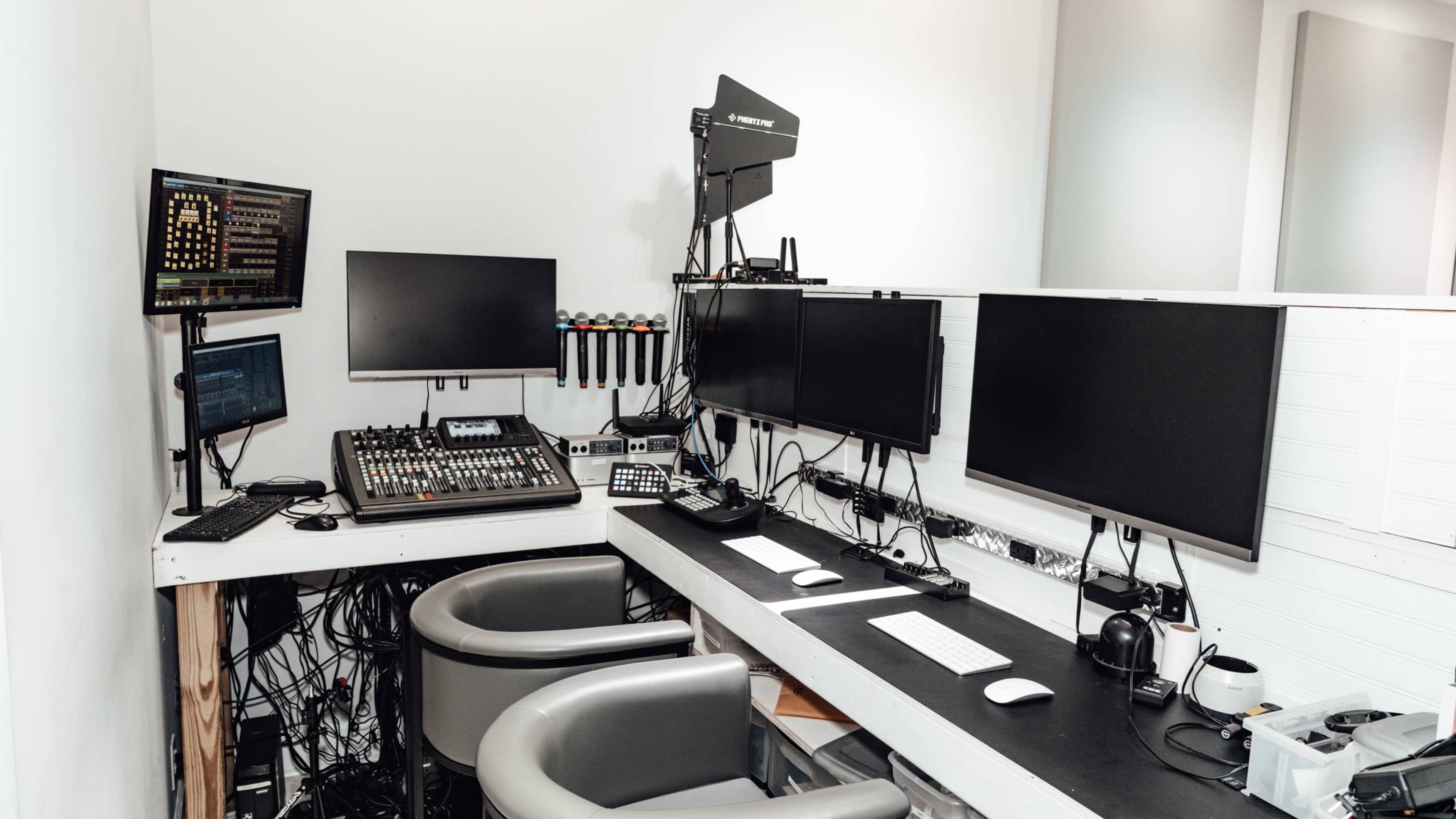 The image shows a modern workspace with multiple computer monitors, audio equipment, and two ergonomic chairs set against a white wall.