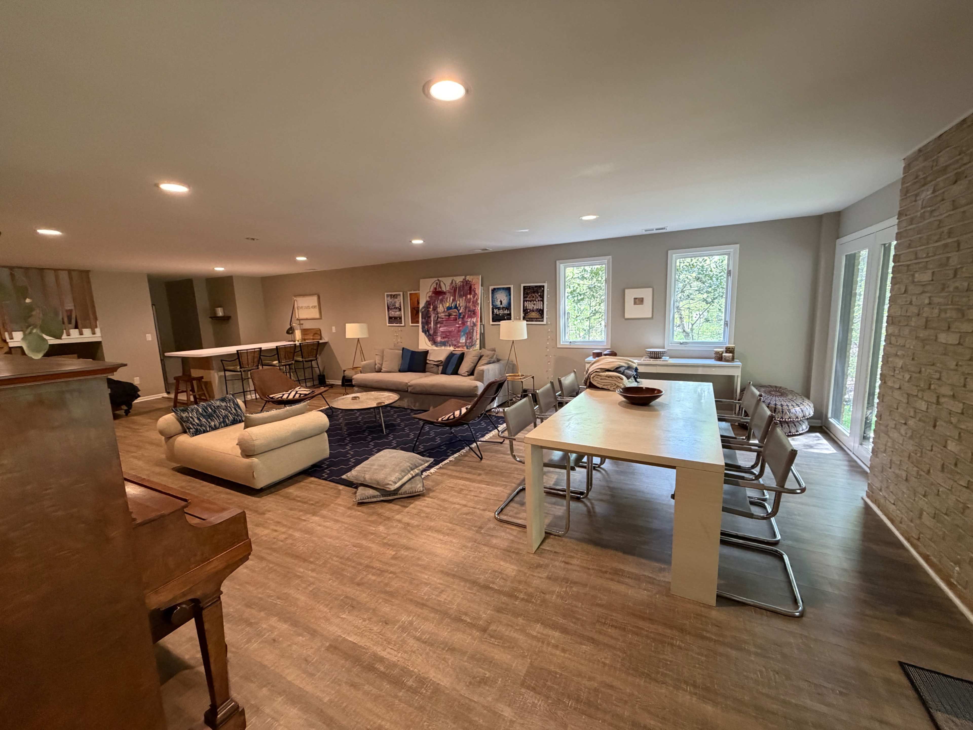 The image depicts a spacious living area with a seating arrangement, dining table, and large windows allowing natural light to enter.