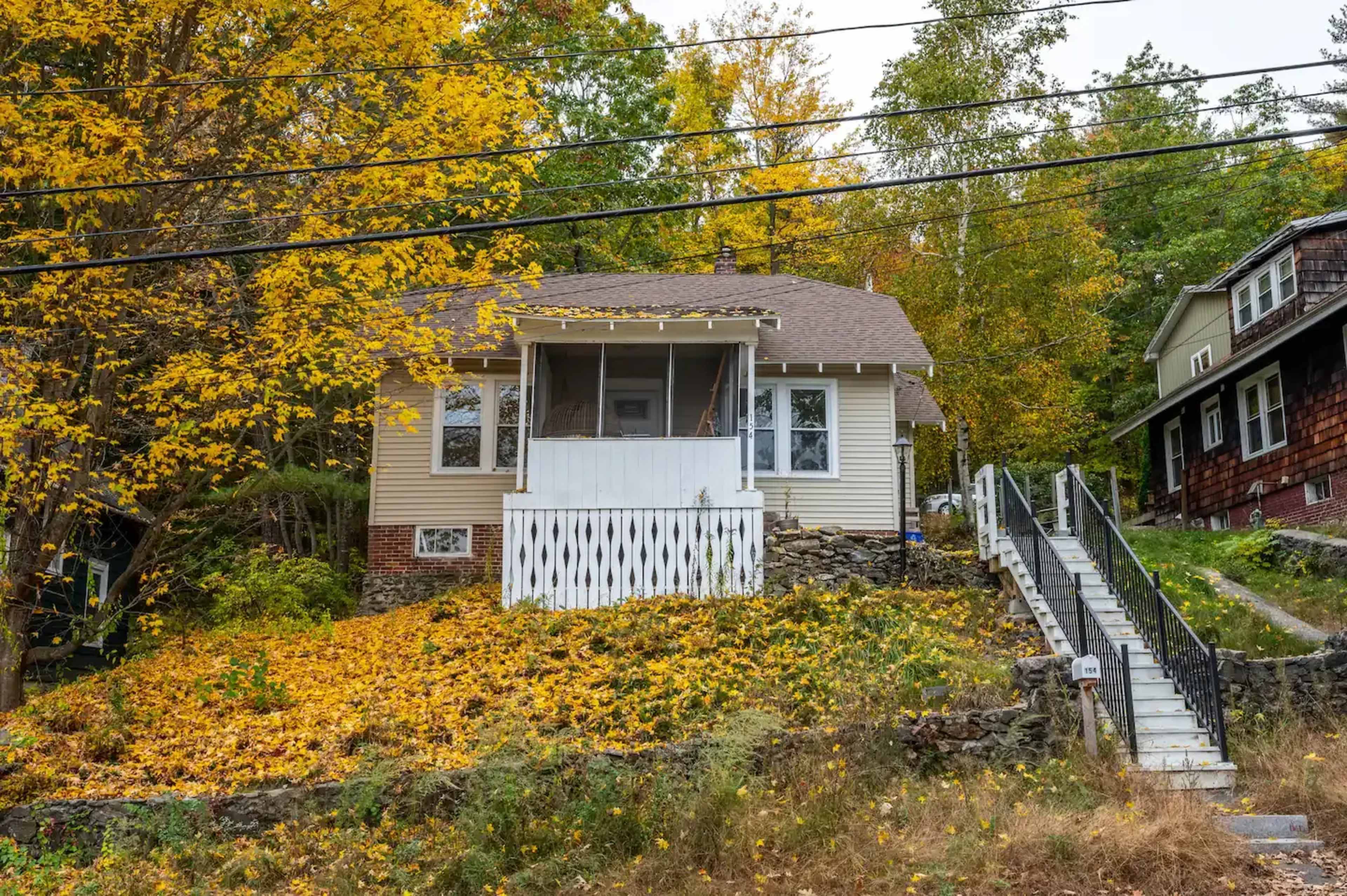 A single-story house with a porch is surrounded by fallen yellow leaves and is situated on a hillside with a set of stairs leading up to it.