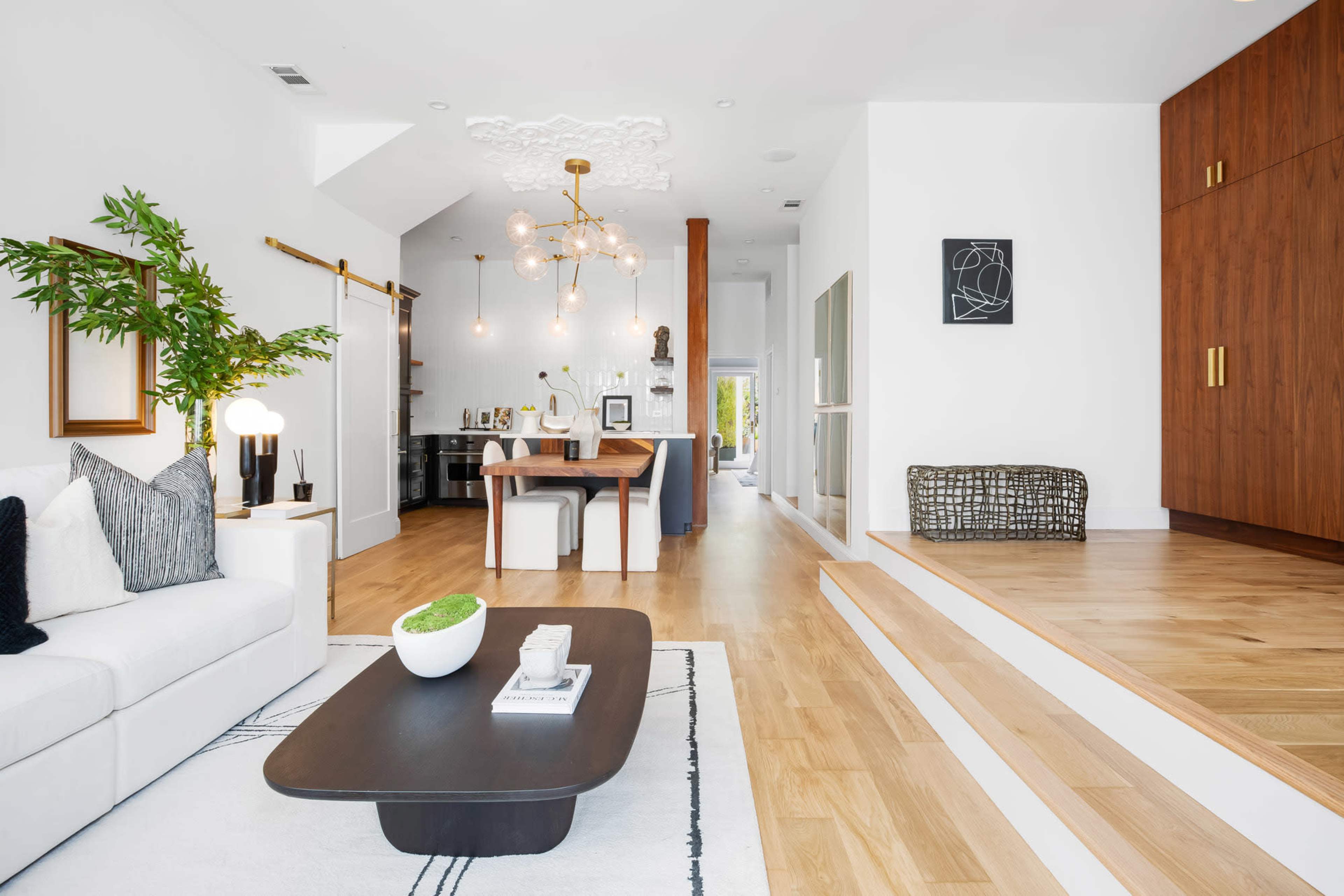 The image shows a modern, open-concept living space featuring a light-filled area with a sofa, decorative plants, a dining table, and a stylish wooden cabinet.