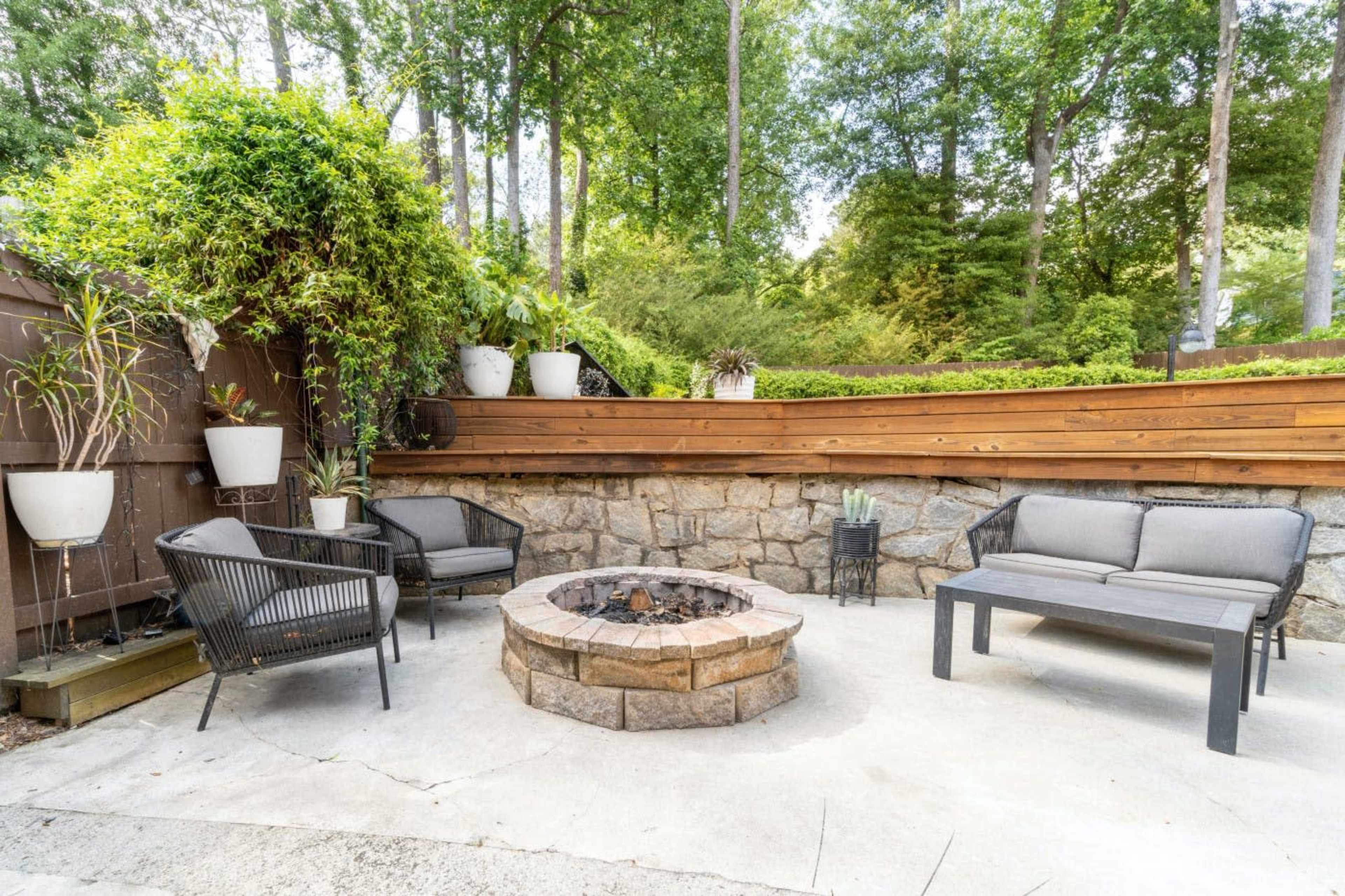 A patio area with a stone fire pit surrounded by chairs and potted plants, set against a backdrop of trees.