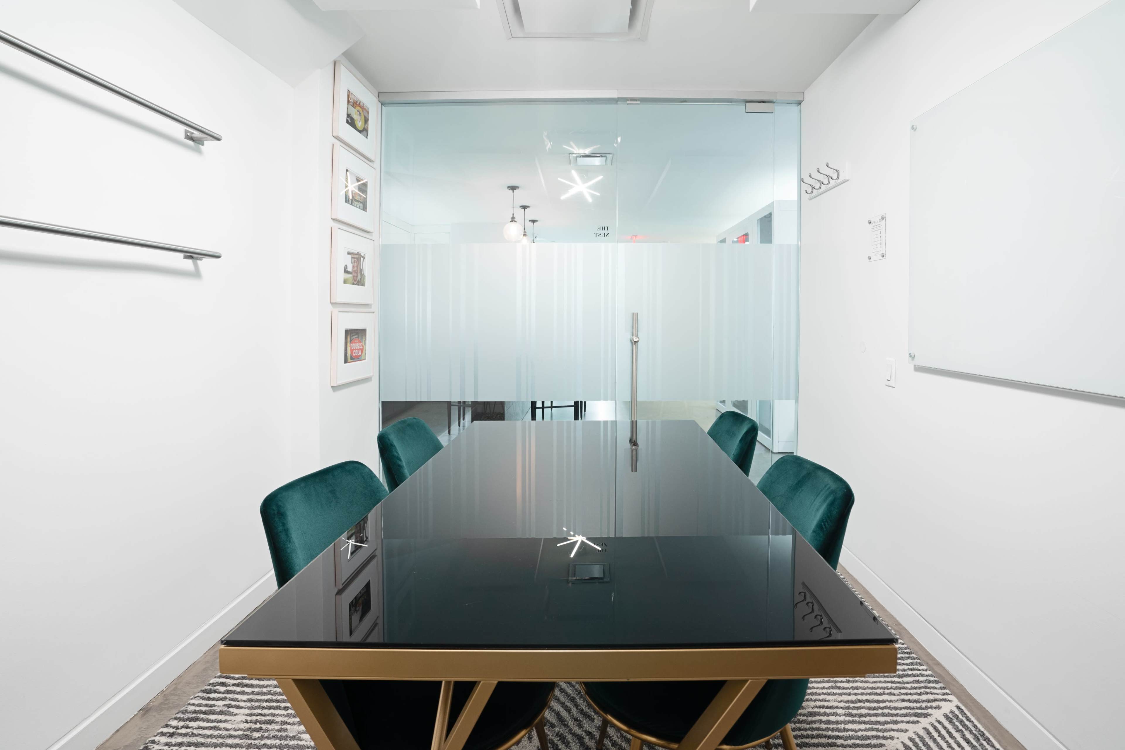 A conference room features a black table surrounded by four green chairs, with glass doors and wall-mounted frames in the background.