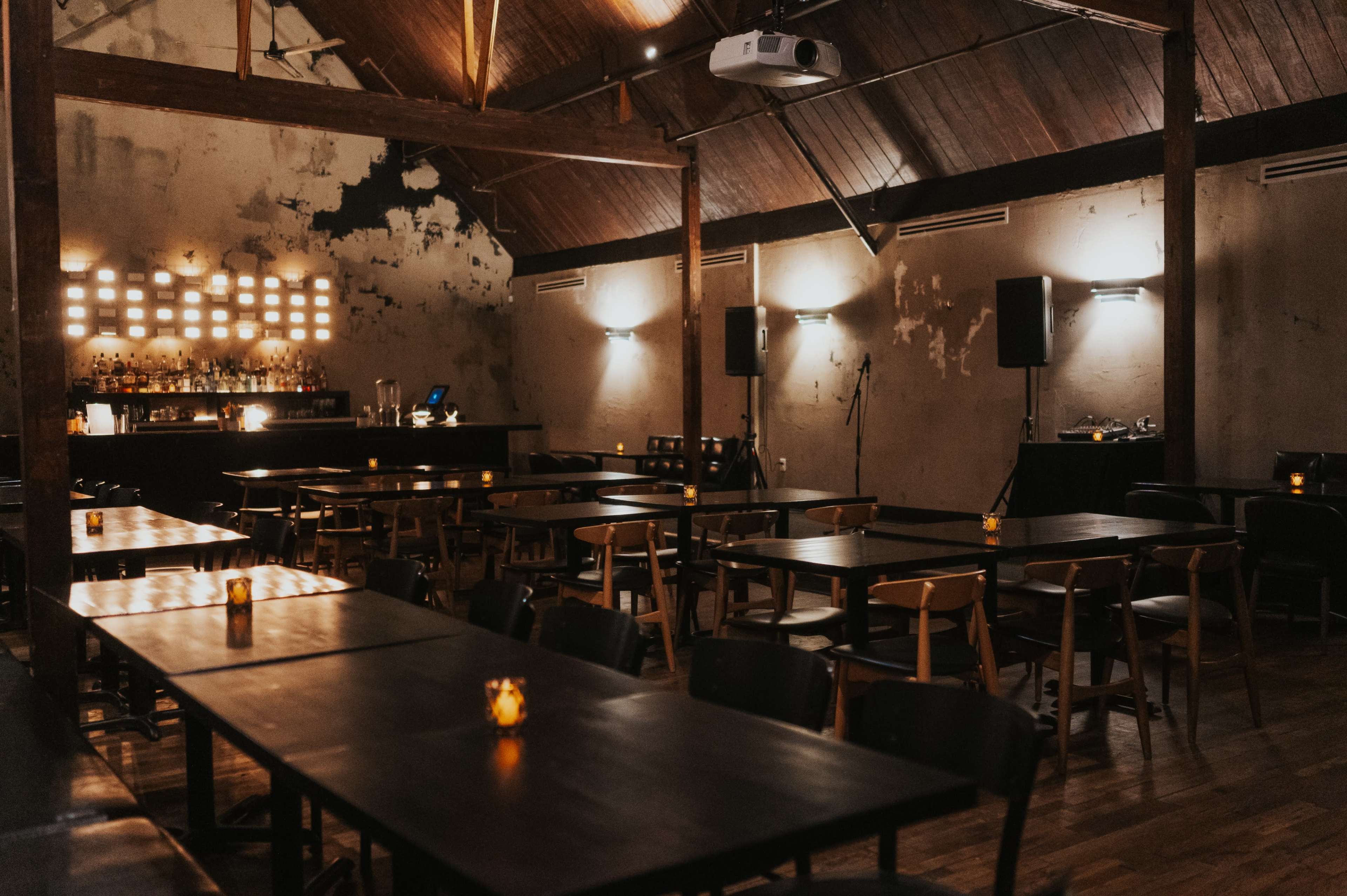 The image shows an empty restaurant or bar with wooden tables, dim lighting, and a bar area at the back.