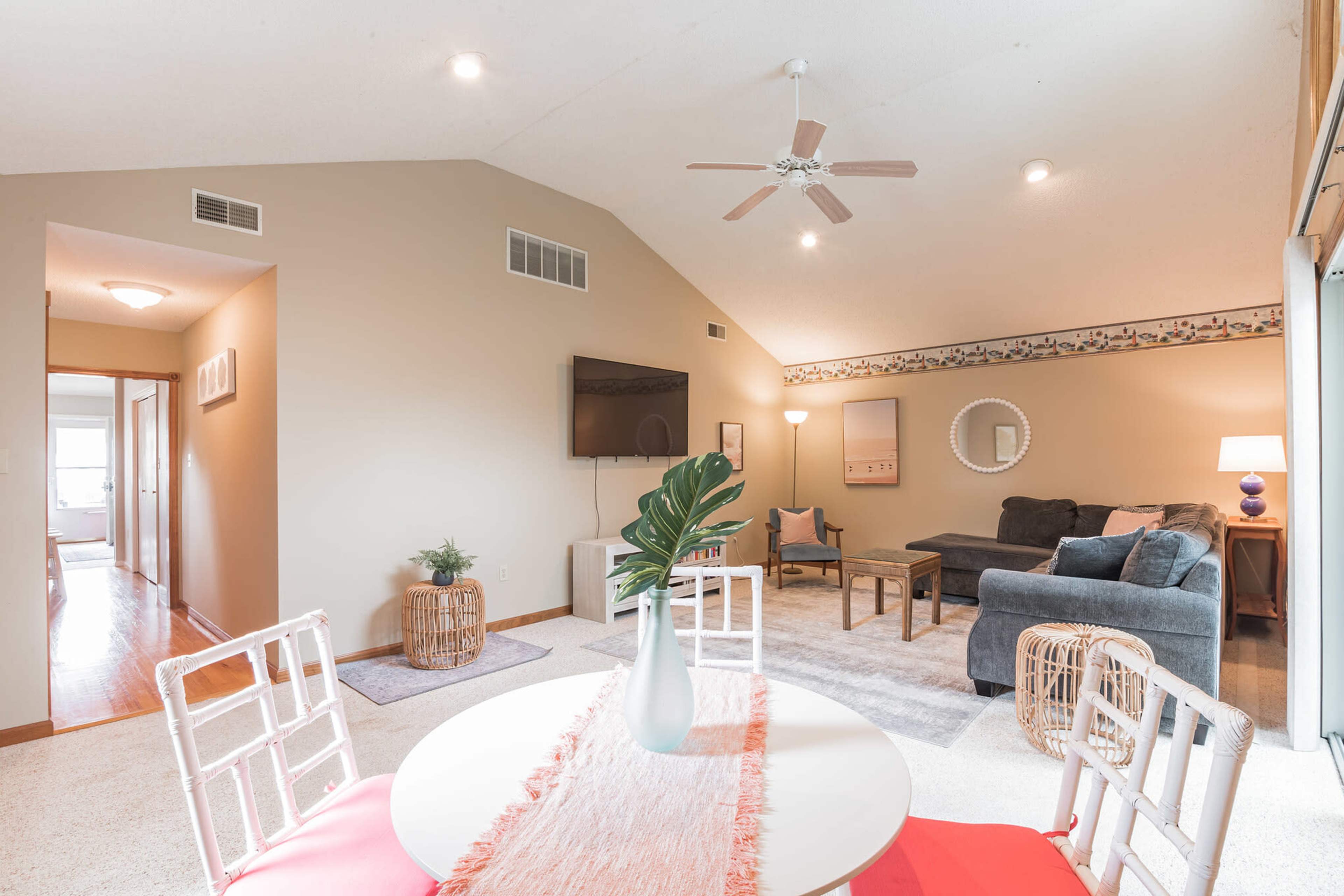 A bright living area with a vaulted ceiling, featuring a dining table with red chairs, a plush sofa, a TV mounted on the wall, and decorative elements.