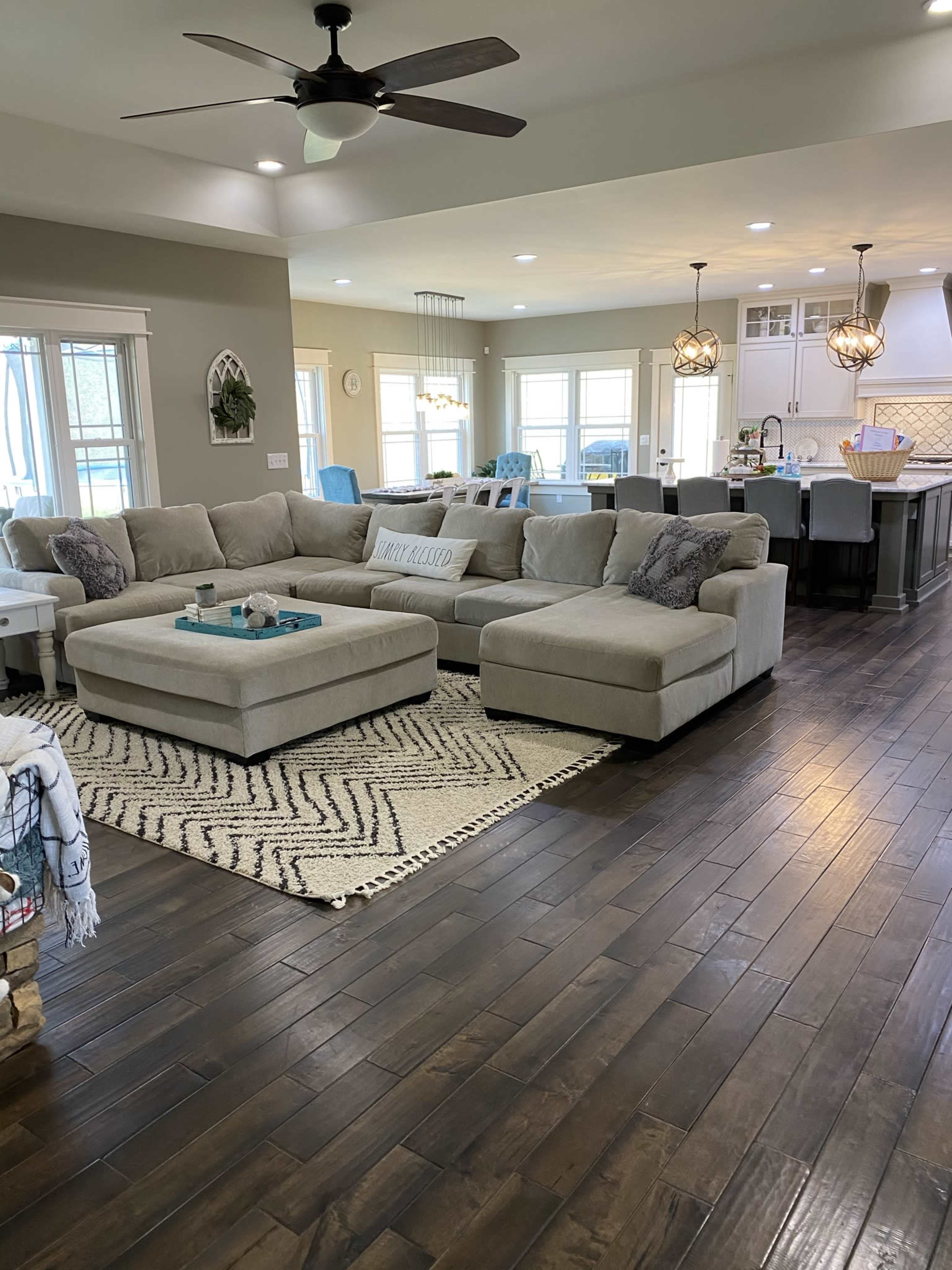 A spacious living room with a large beige sectional sofa and a patterned area rug, leading to an open kitchen area with modern fixtures and large windows.
