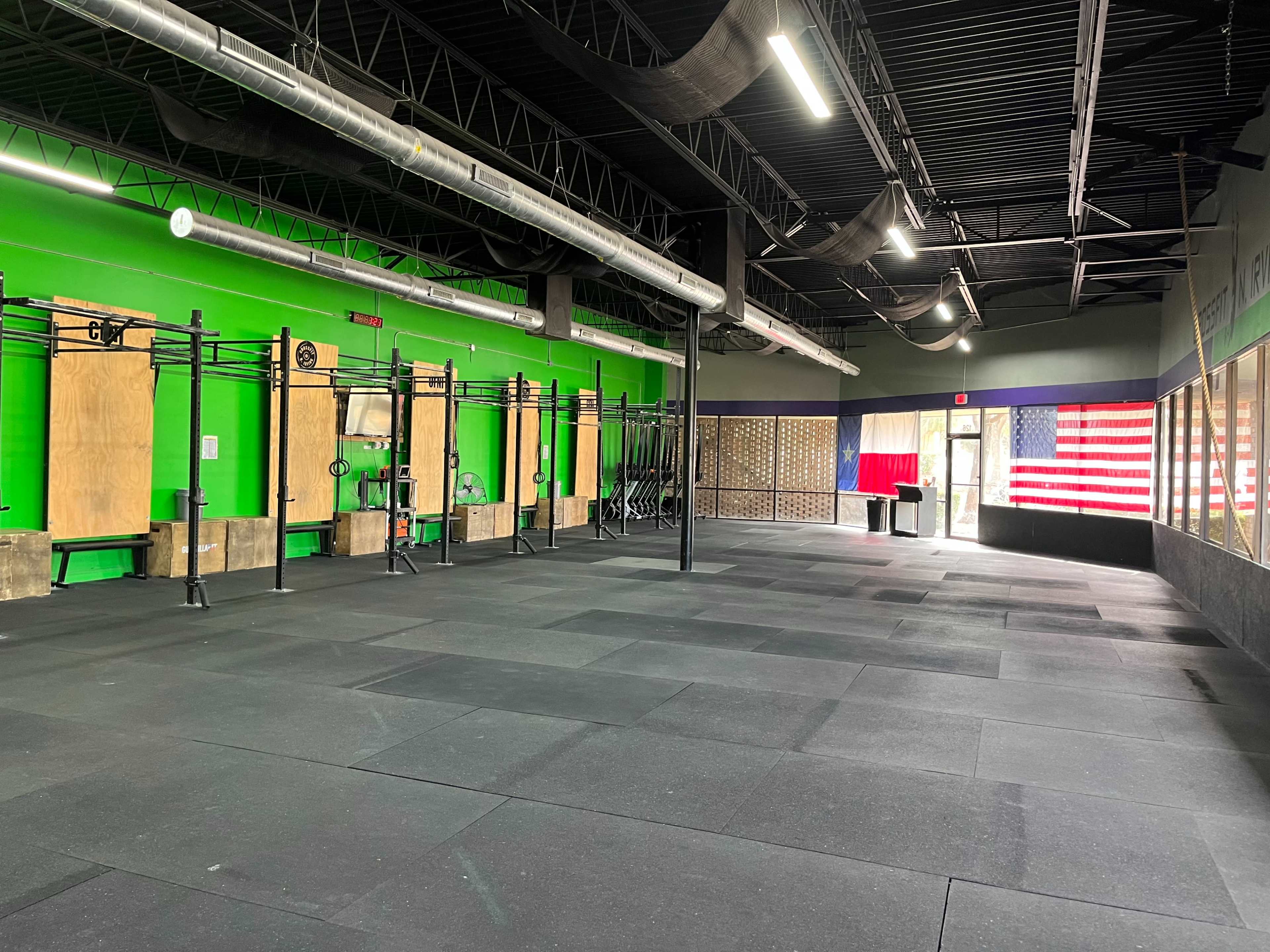 An indoor gym space featuring black rubber flooring, green walls, and various gym equipment arranged along one side.