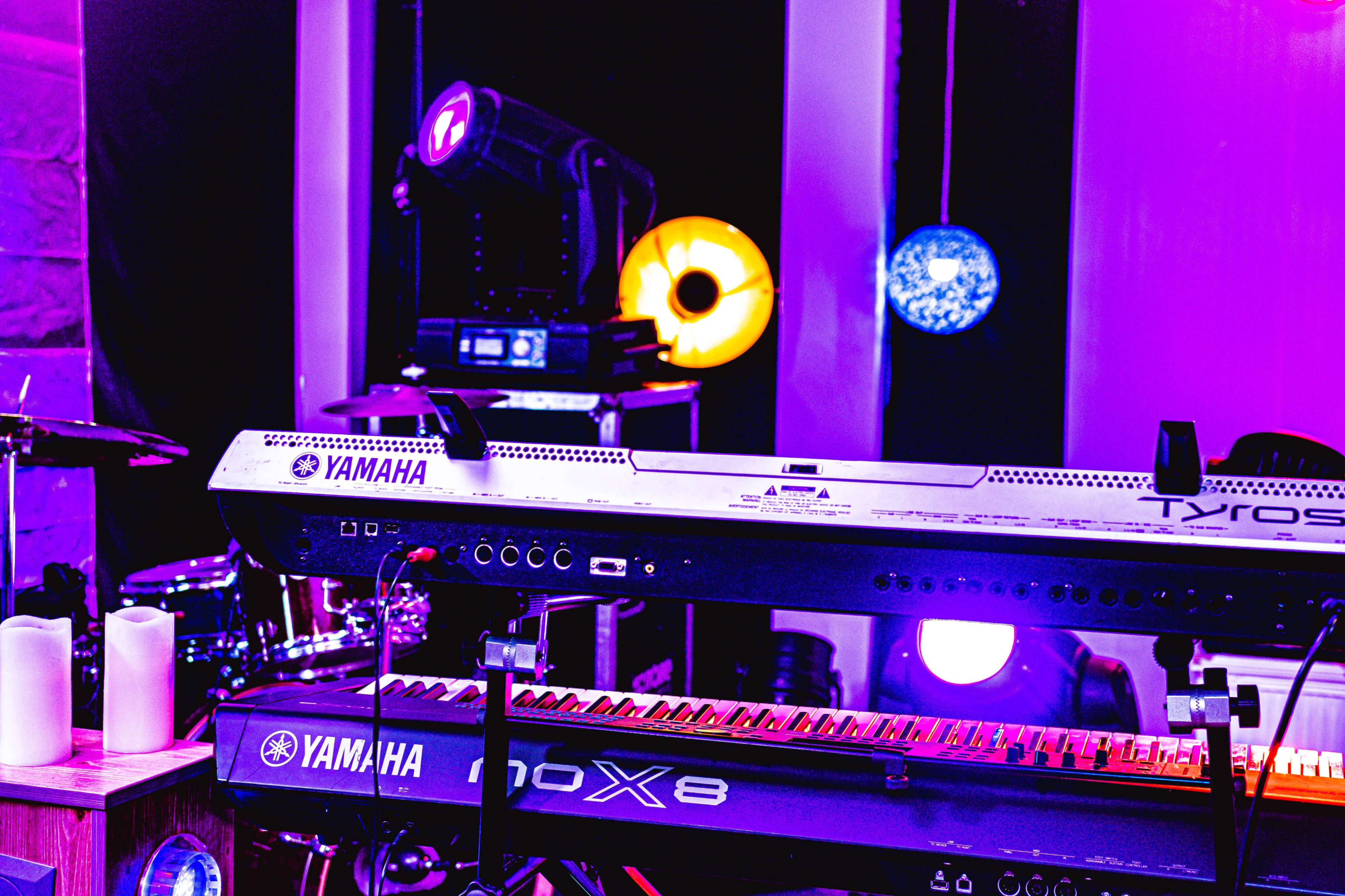 A Yamaha keyboard is positioned in front of a drum set and illuminated by colorful stage lights.