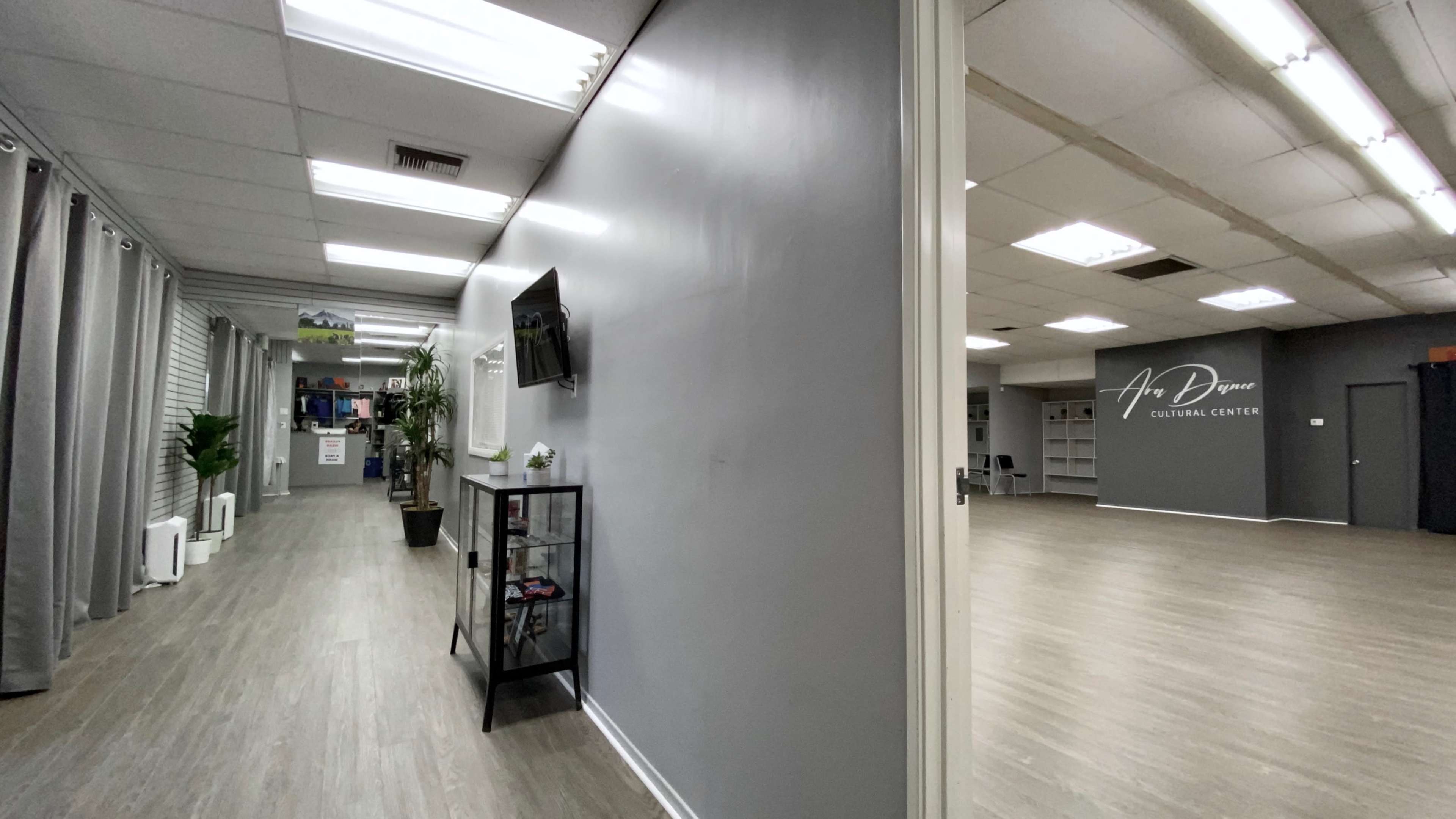 A spacious cultural center featuring a bright, open room on the right and a hallway with a wall-mounted TV and plants on the left.