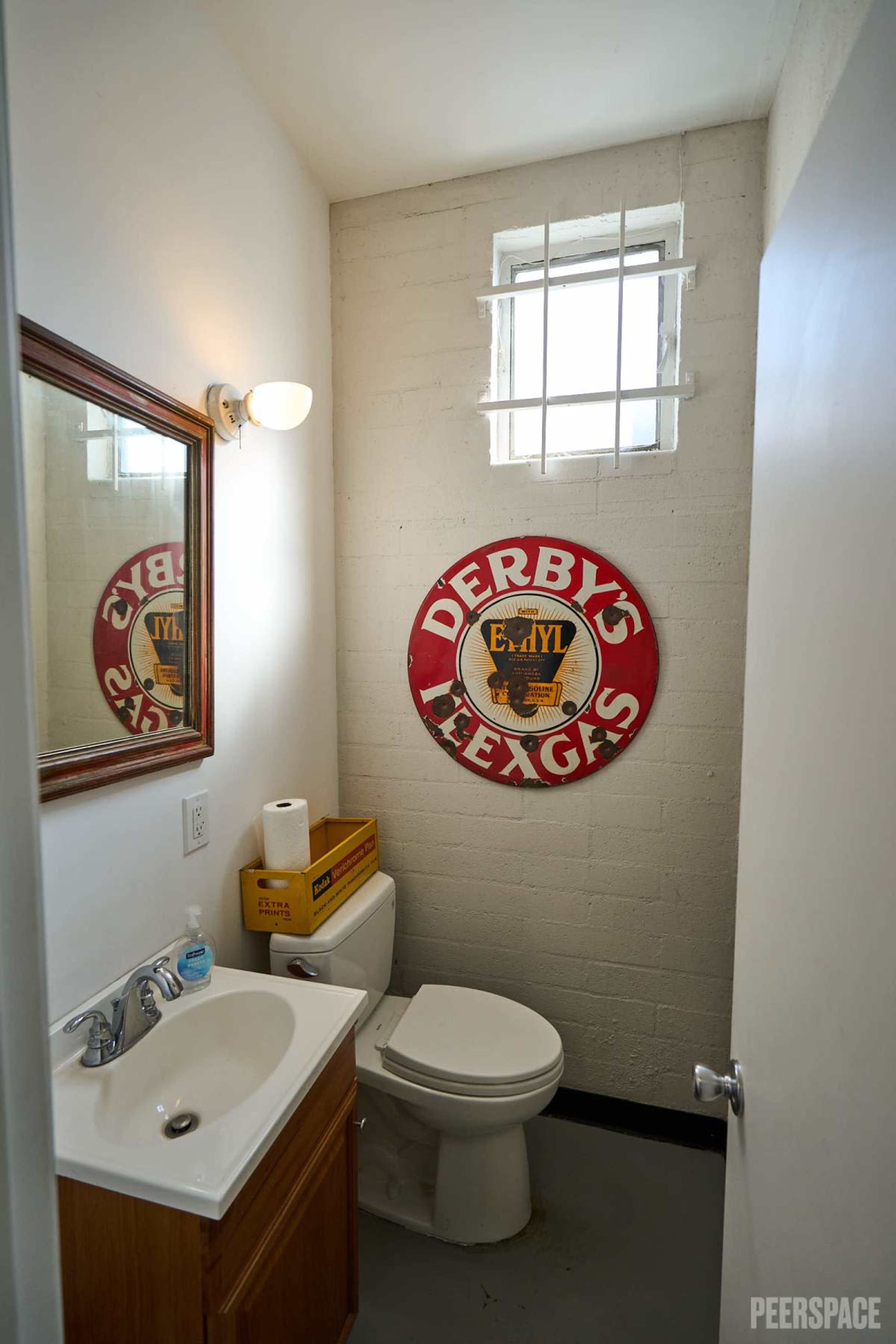 The image shows a small bathroom featuring a white toilet, a sink with a wooden cabinet, a mirror, and a vintage sign on the wall.