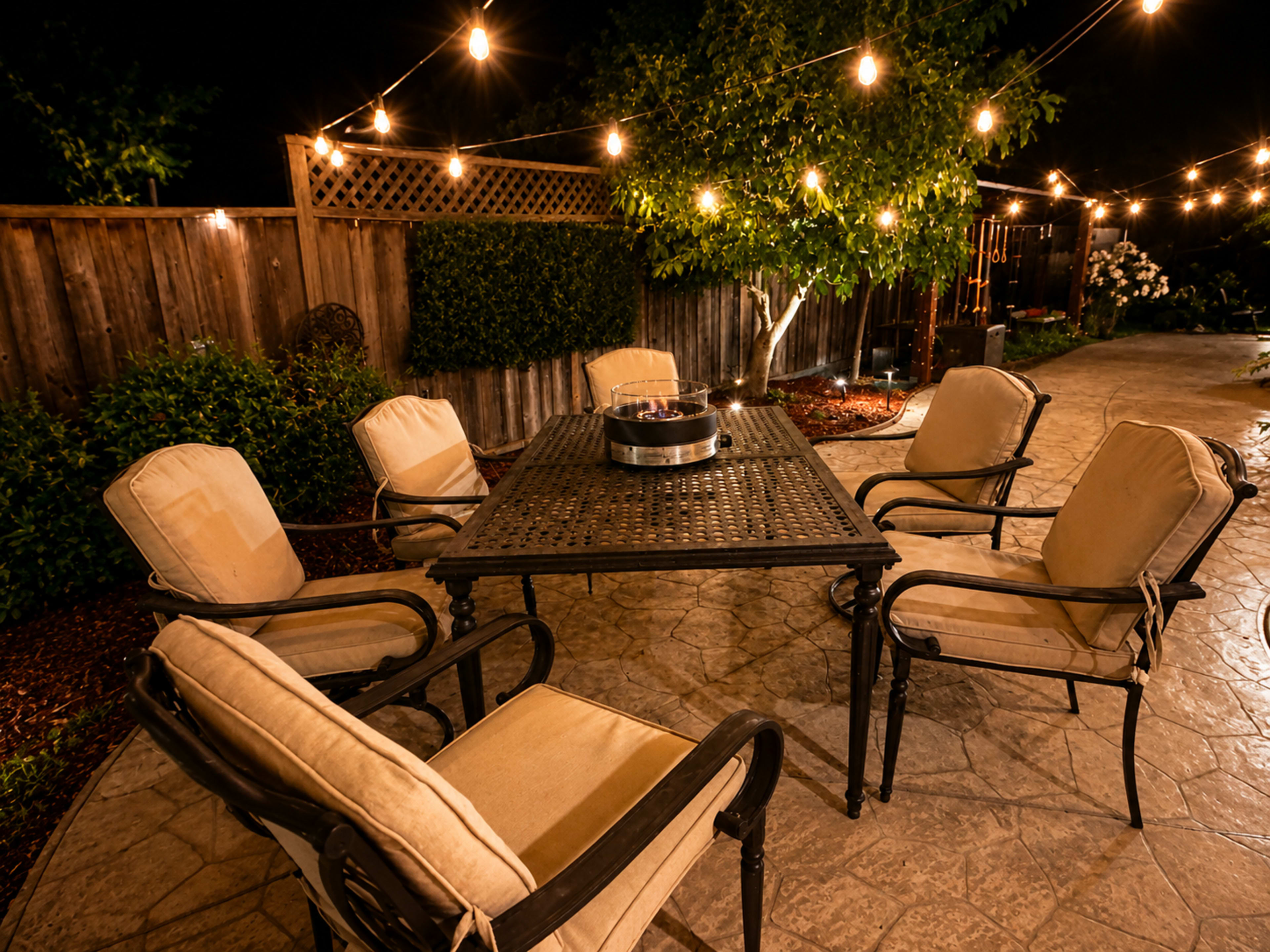 A backyard patio features a table surrounded by six chairs and illuminated by string lights overhead.