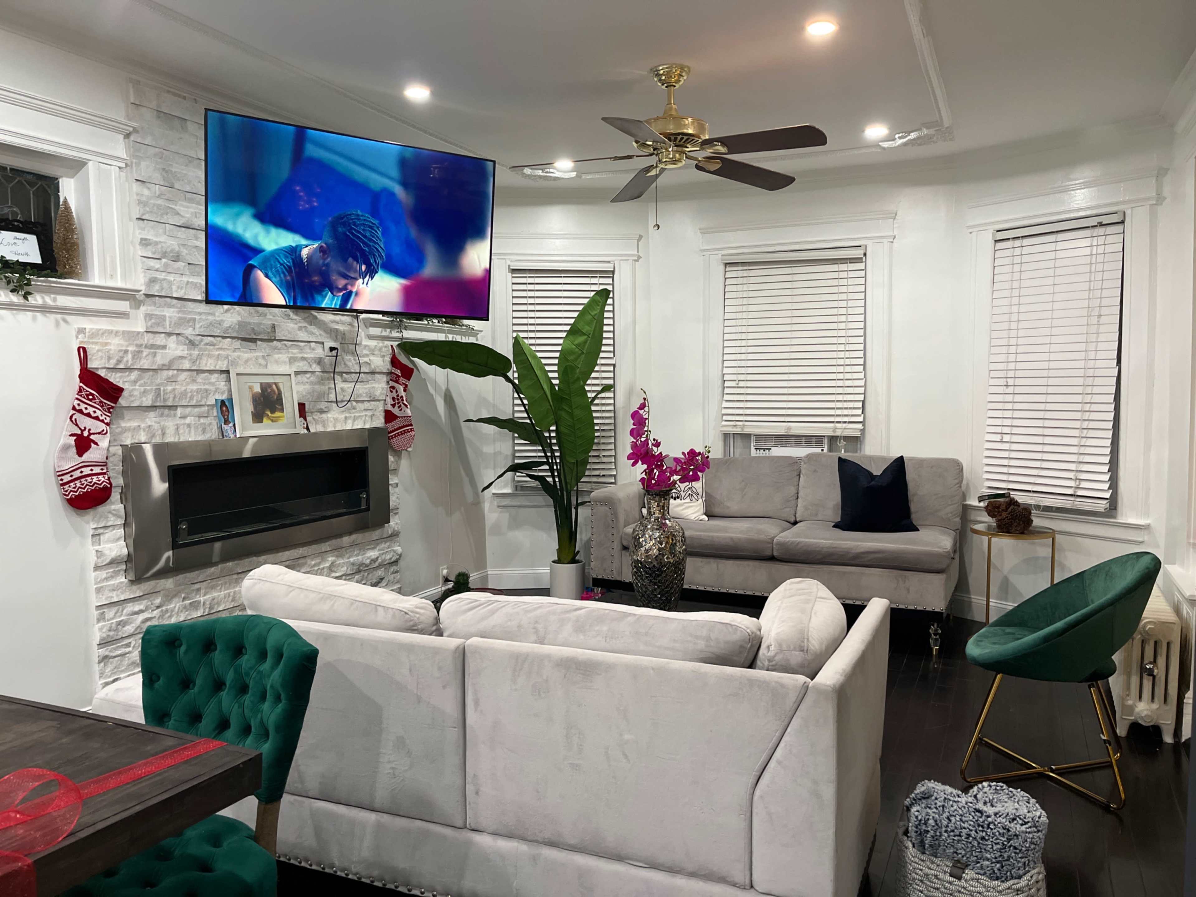 A living room features a television mounted on the wall, a modern gray sofa, a green accent chair, a decorative plant, and a fireplace with stockings hung nearby.