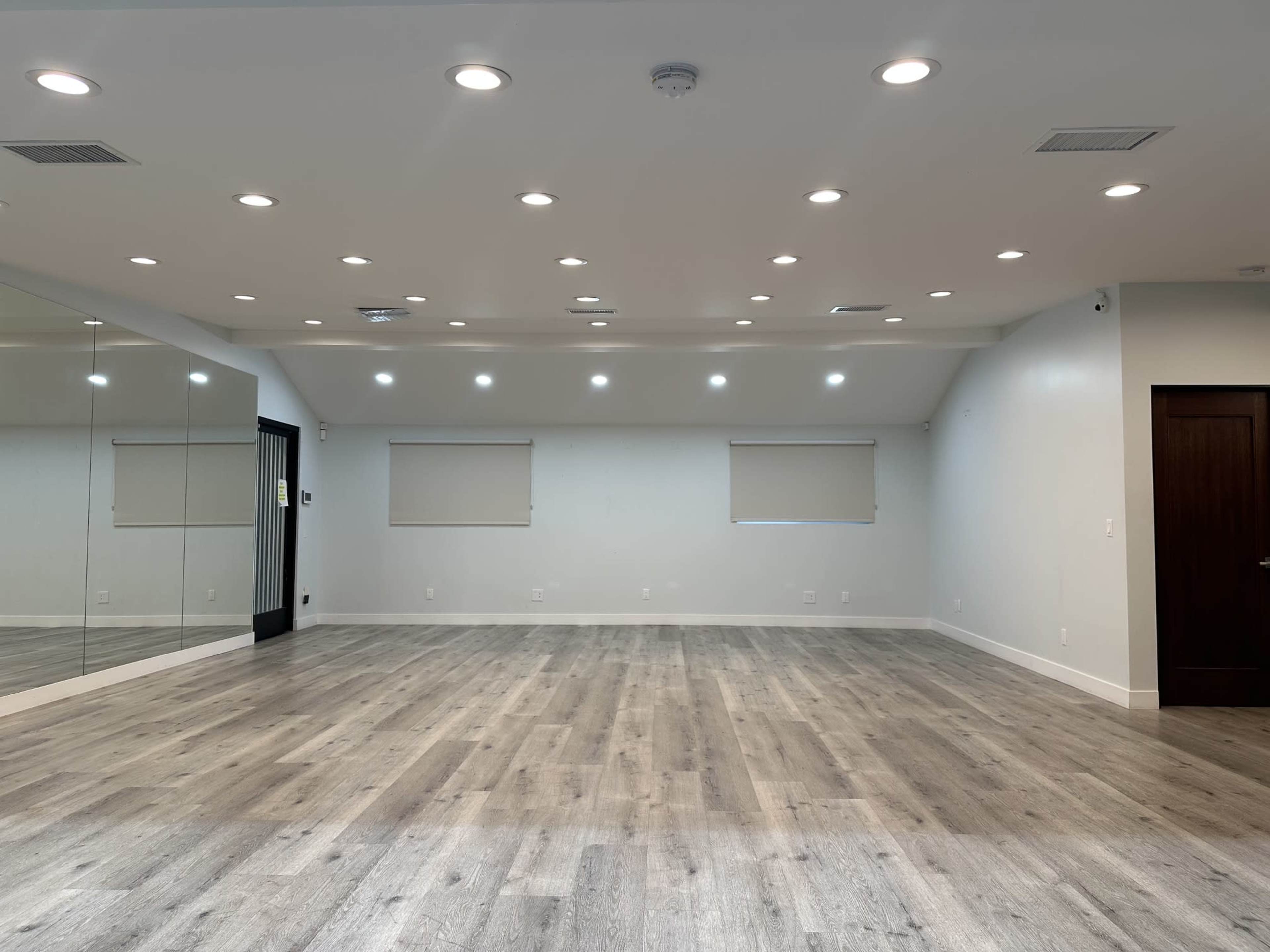 The image shows an empty room with wooden flooring, recessed lighting, and mirrors on one wall.