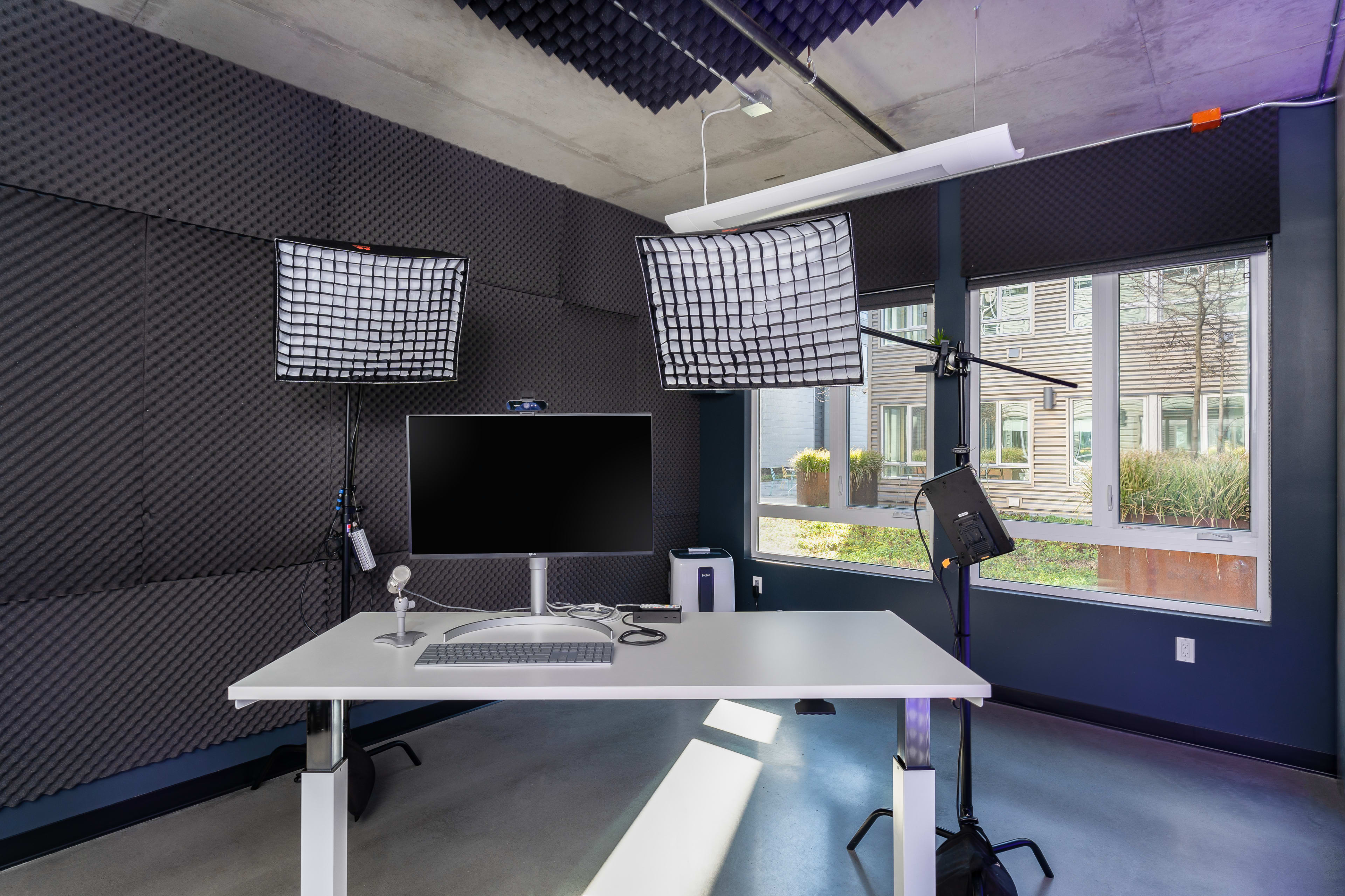 A modern recording studio with a white desk, computer monitor, and professional lighting setup against a backdrop of soundproofing panels.