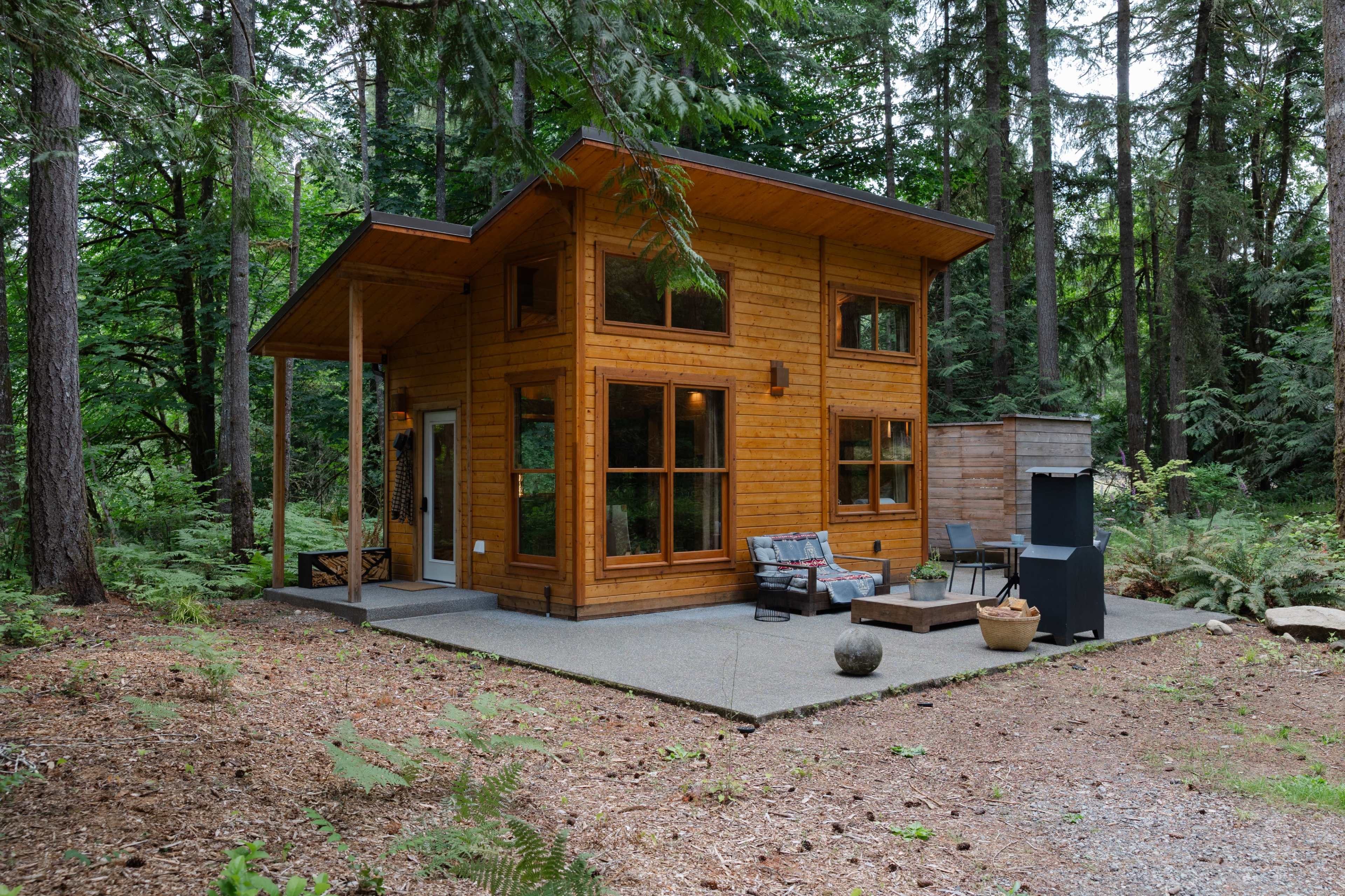 A small, wooden cabin with large windows is situated in a forested area surrounded by trees and greenery.