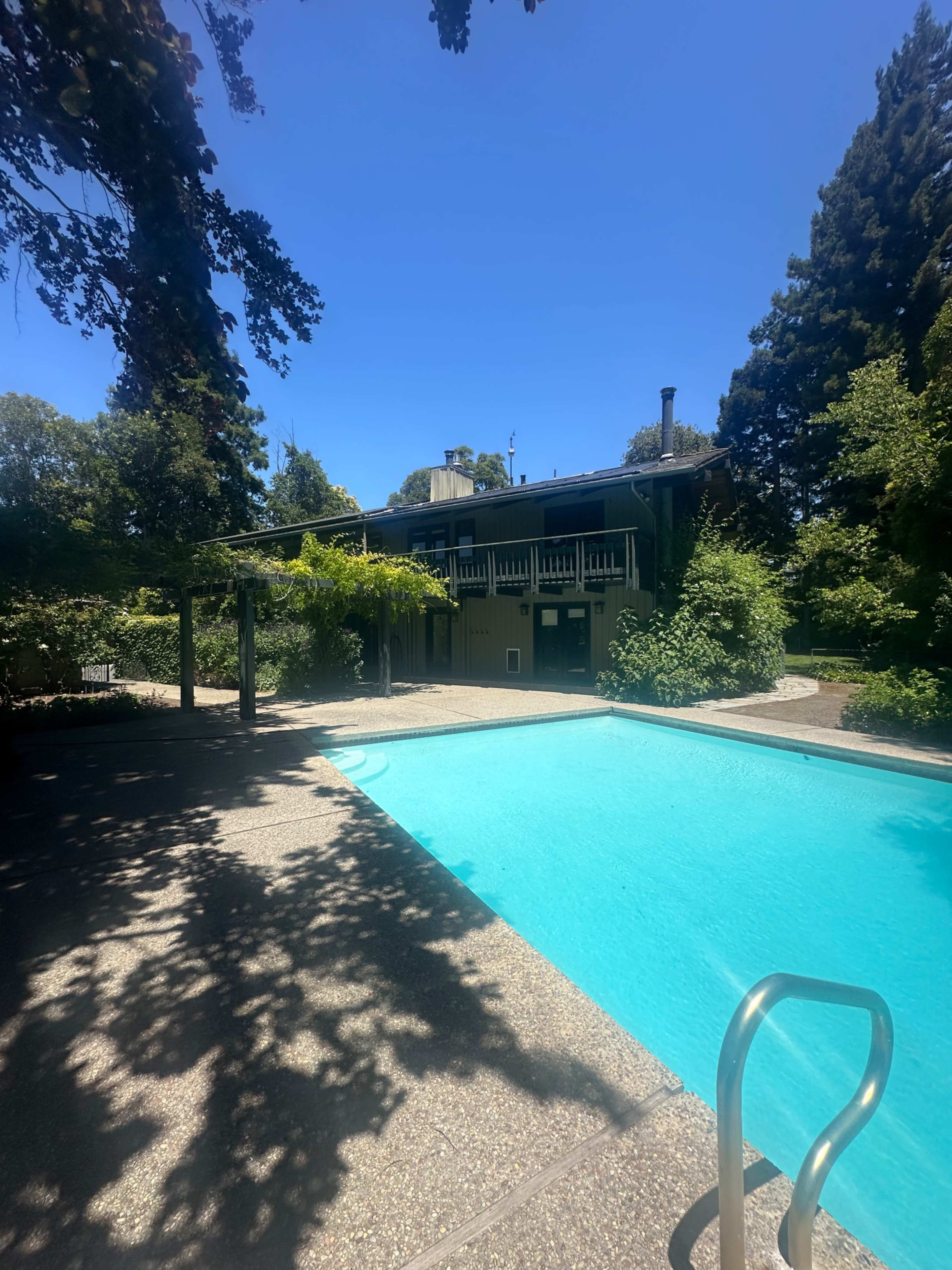A clear blue swimming pool is situated in front of a two-story house surrounded by tall trees and lush greenery.