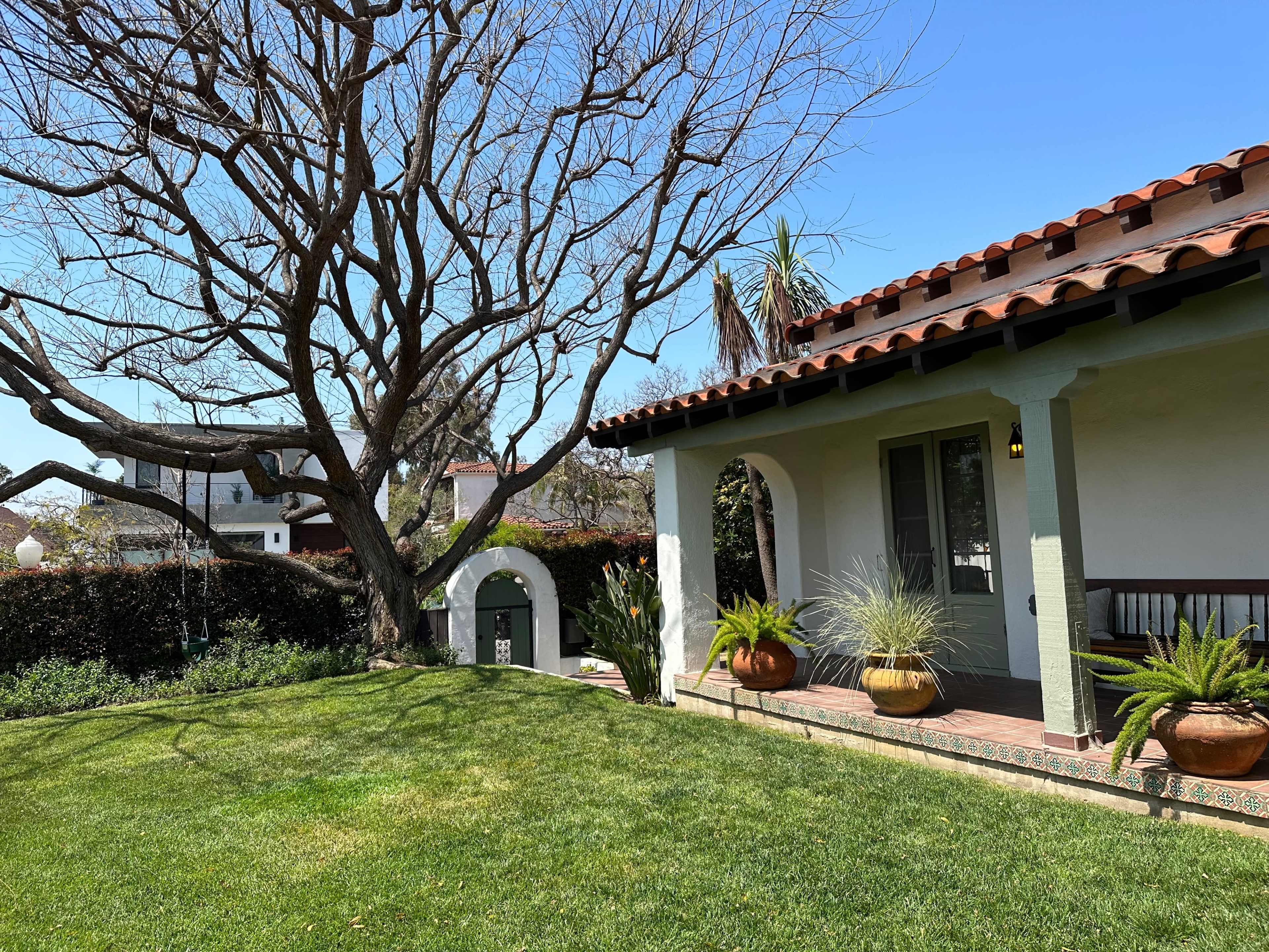 A house with a tiled roof and a porch is surrounded by a well-maintained lawn and a bare tree, with decorative potted plants positioned nearby.