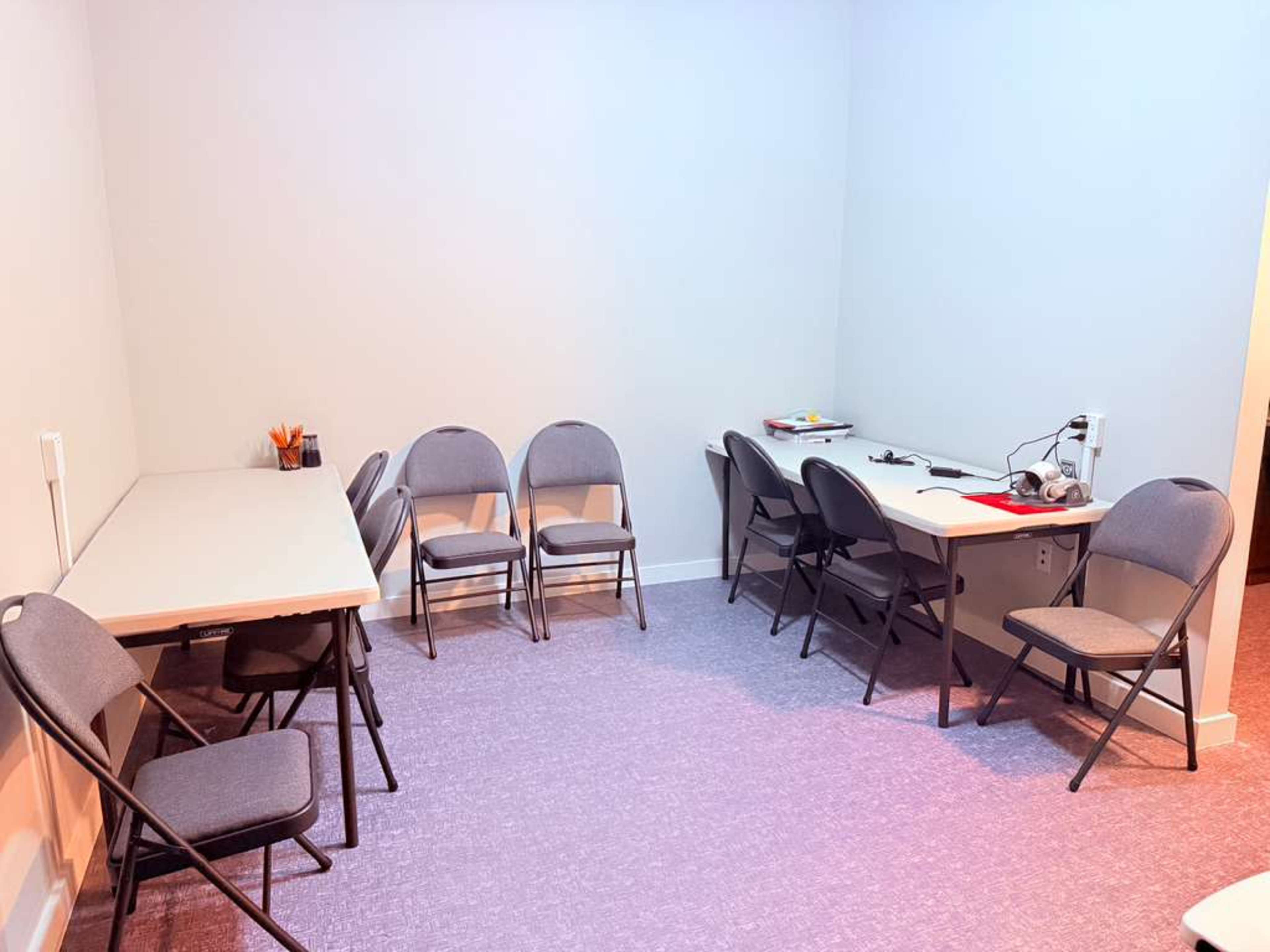 The image shows a small, sparsely furnished room with several folding chairs and two tables, one of which has various items like a computer and headphones on it.