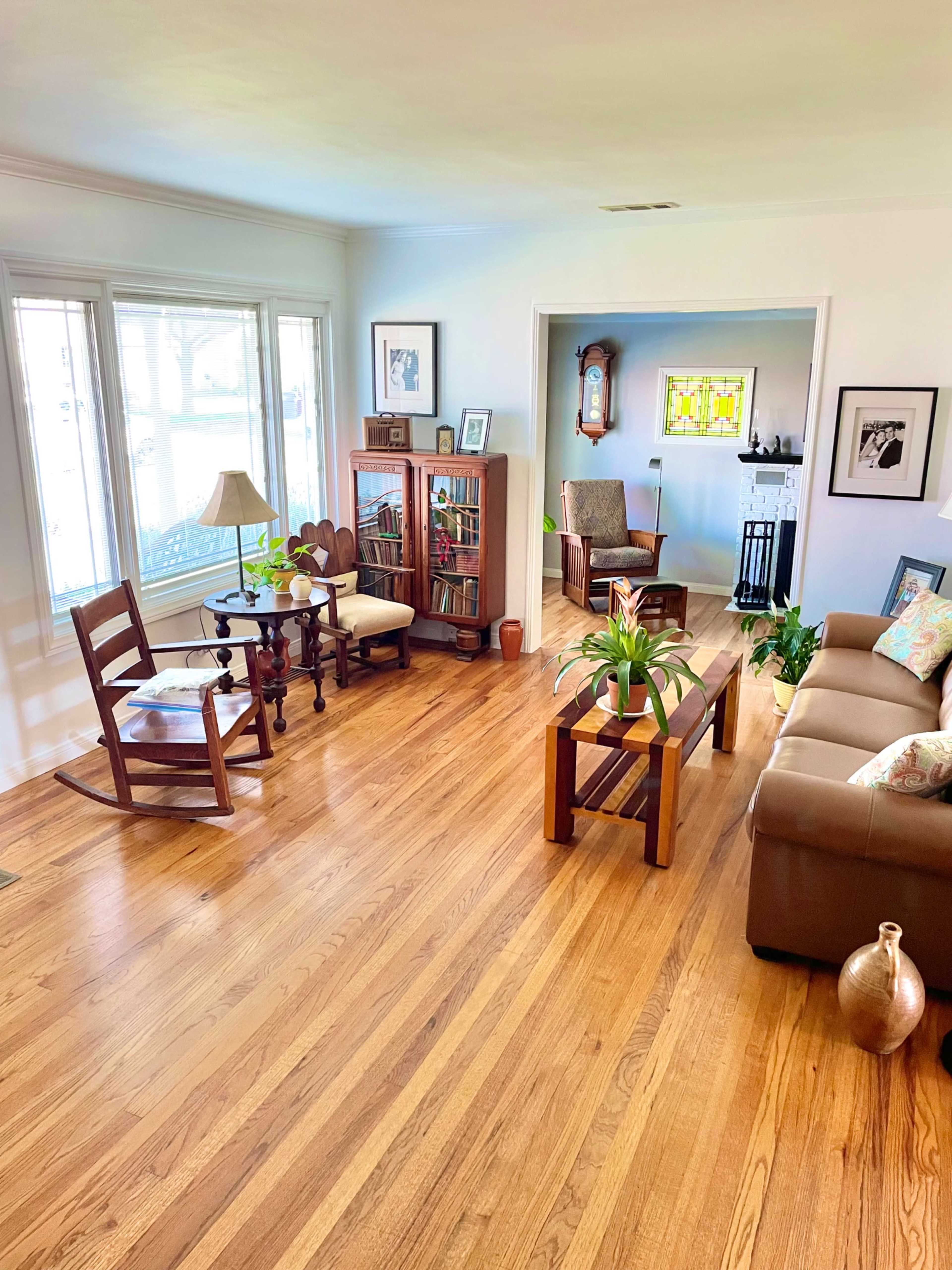 A brightly lit living room with wooden floors, a seating area, a rocking chair, a bookshelf, and plants.