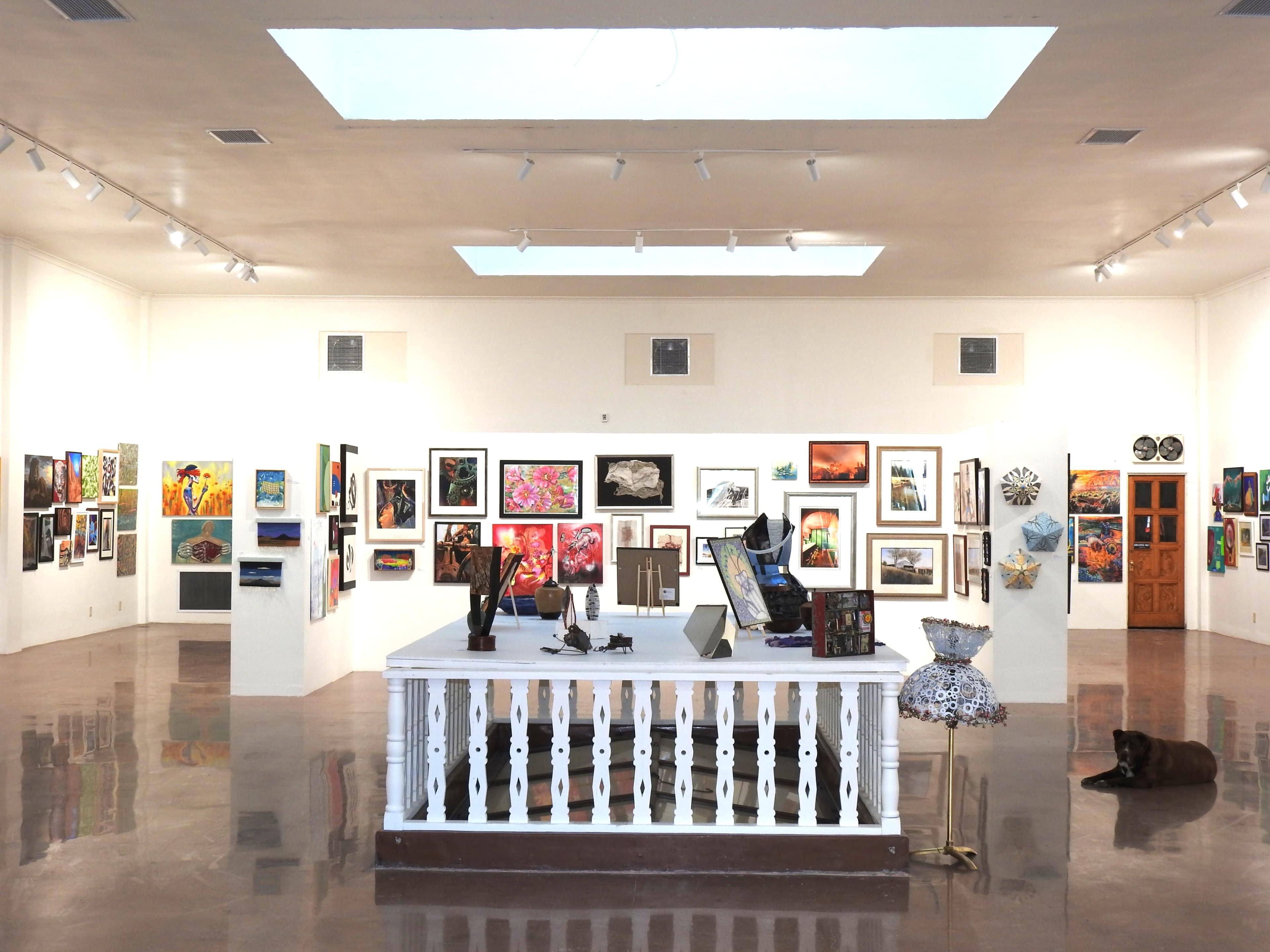 The image shows a brightly lit art gallery with various artworks displayed on the walls and a central white table featuring several art pieces.