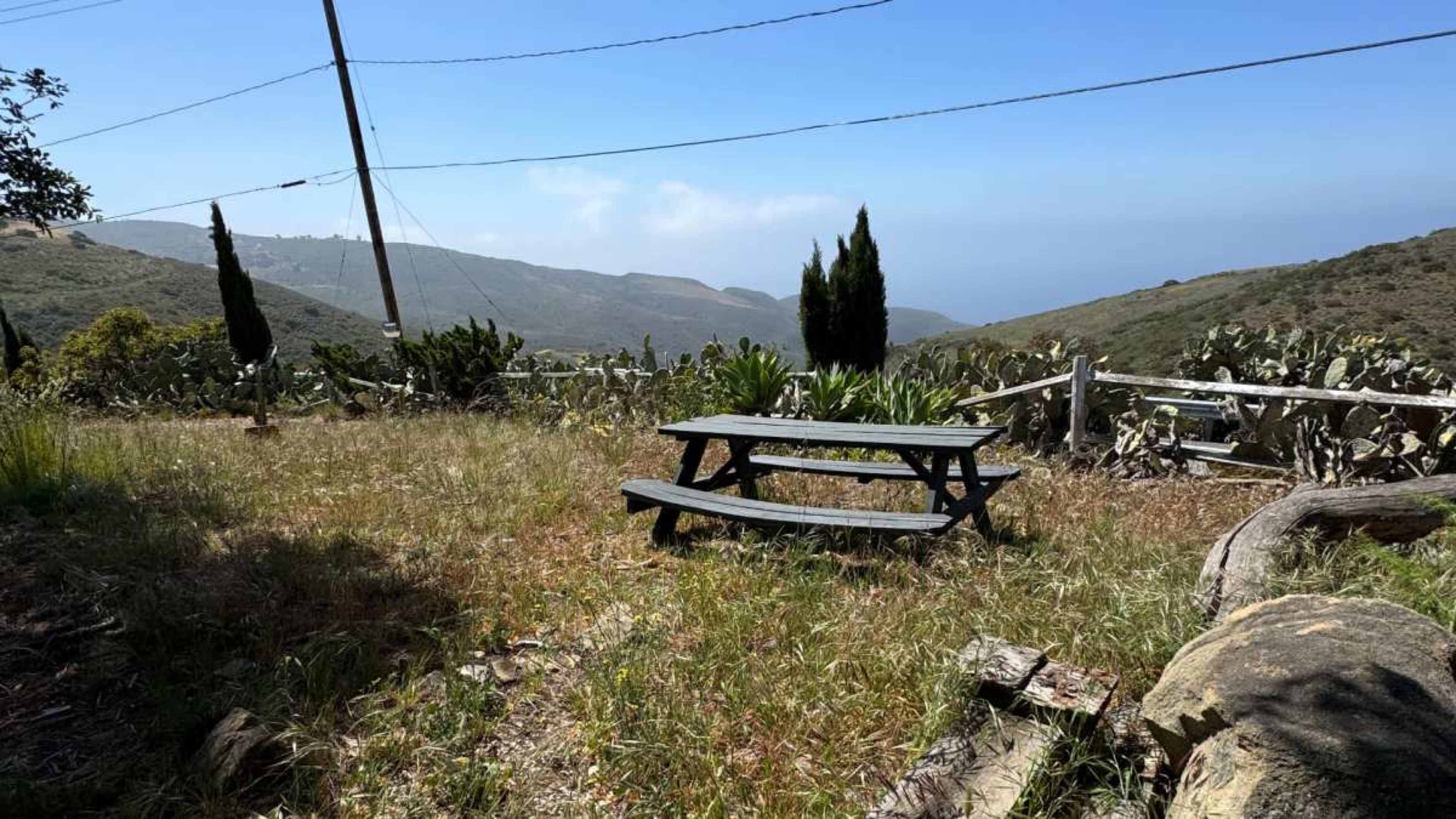 A wooden picnic table sits in a grassy area overlooking a mountainous landscape and the ocean in the distance.