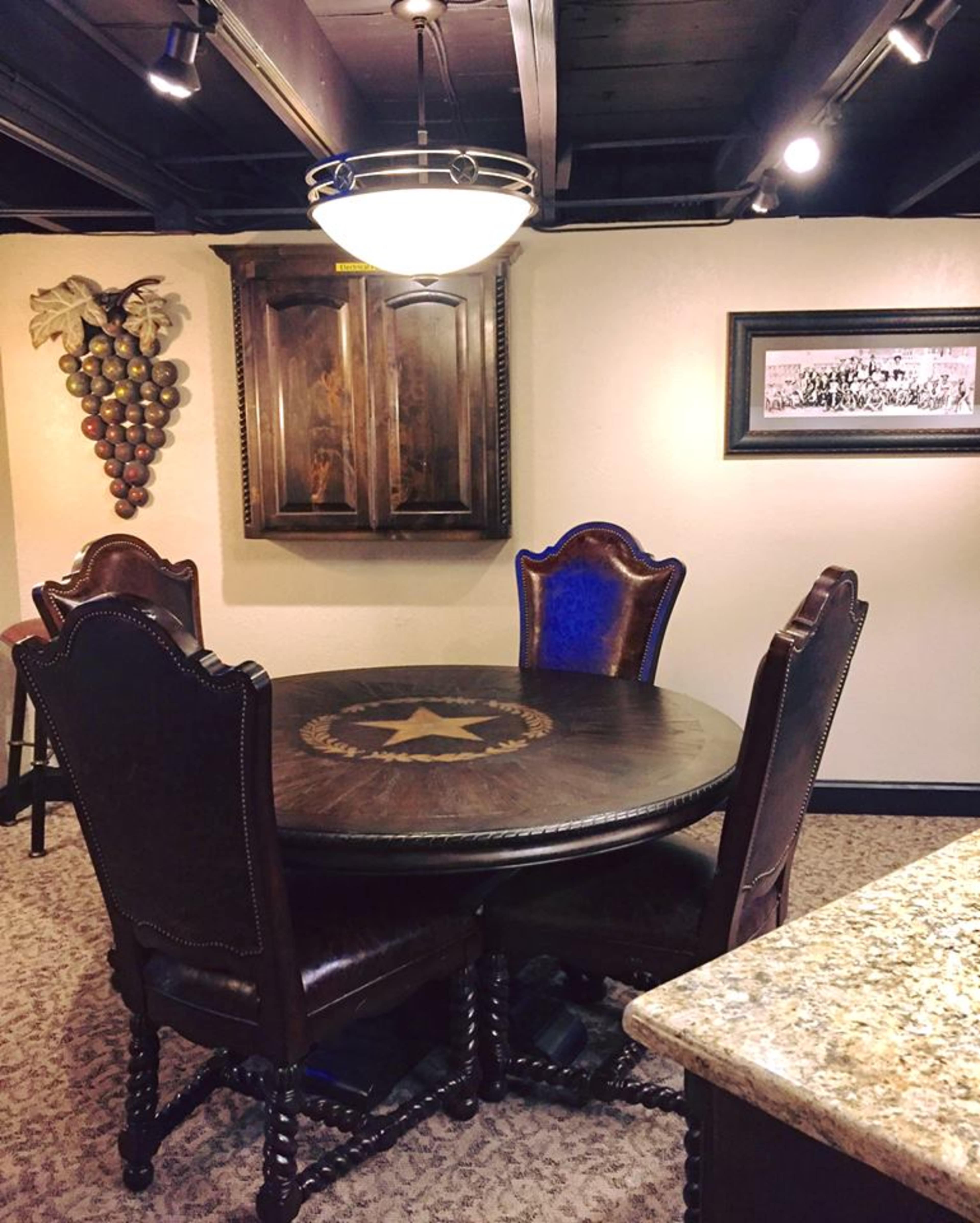 A dining area with a round wooden table and four upholstered chairs, illuminated by a ceiling light, with decorative elements including a grapevine wall decor and a framed picture.