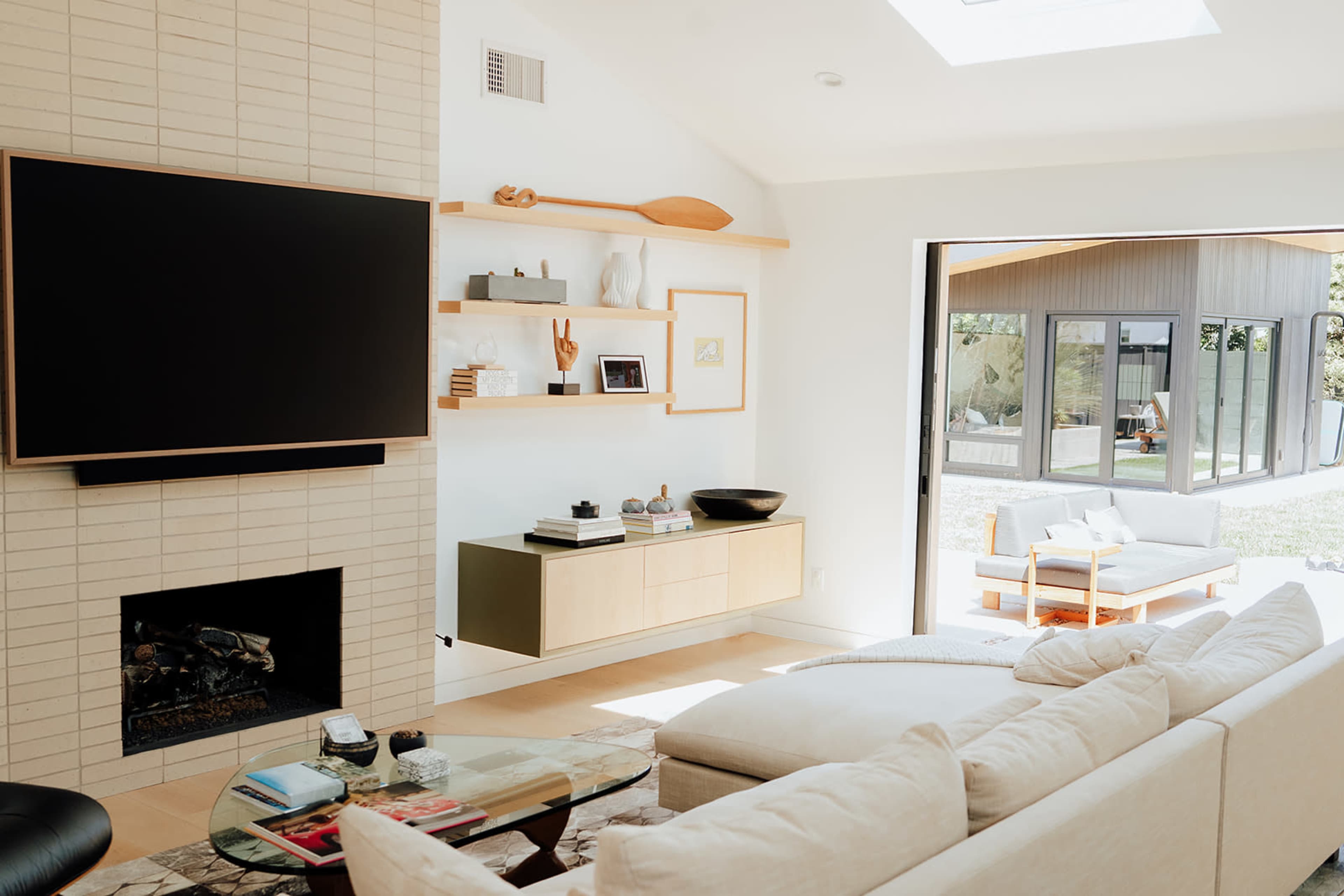 A modern living room features a large flat-screen TV mounted above a sleek fireplace, with light-colored furniture and large windows that open to a lush outdoor space.