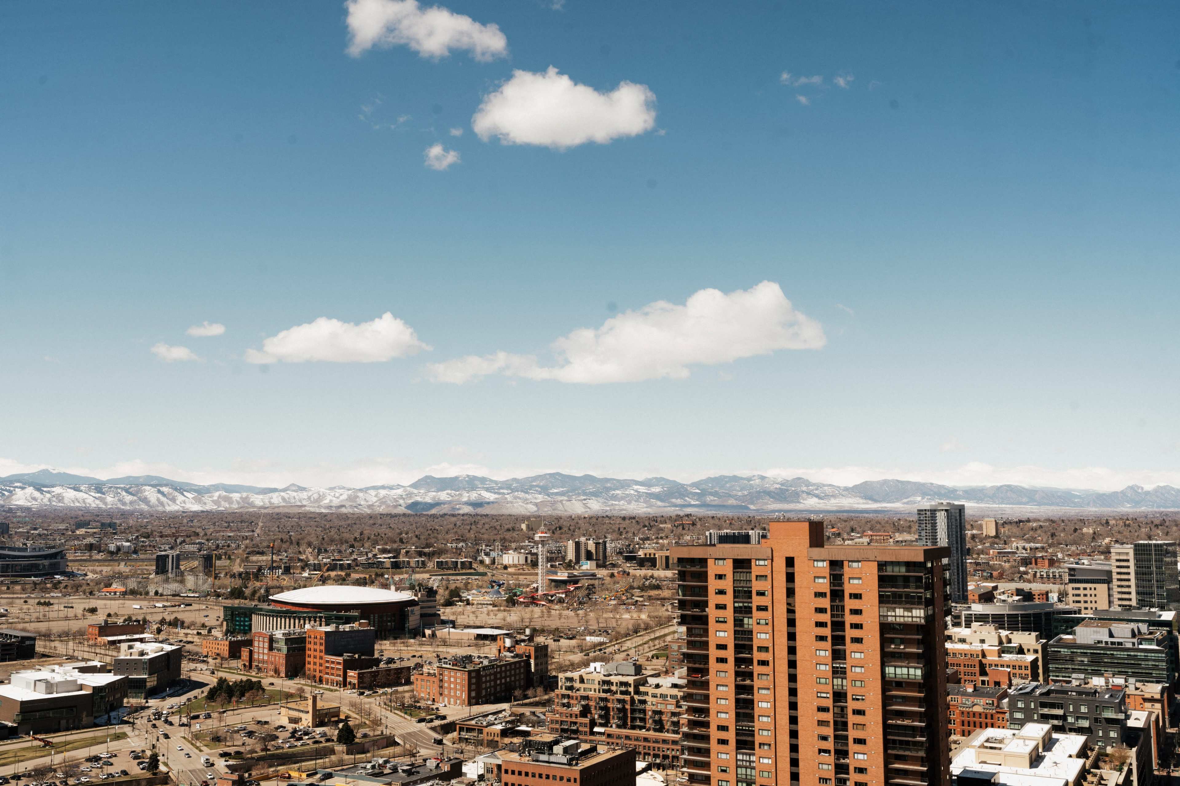 A panoramic view of a city with a mix of modern buildings and distant snow-capped mountains under a clear blue sky.