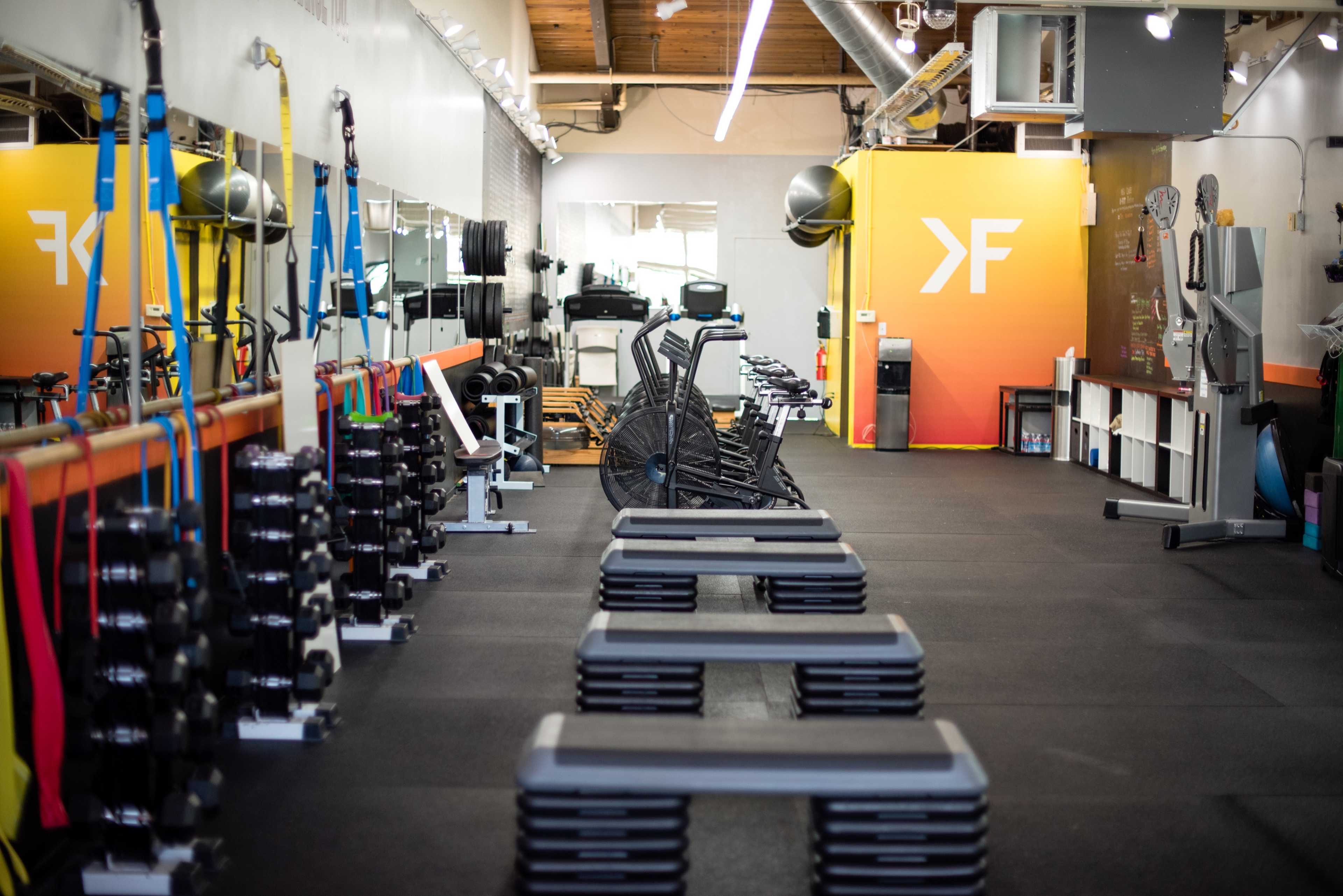 The image shows a well-equipped gym featuring exercise machines, free weights, and step platforms arranged in an organized manner.