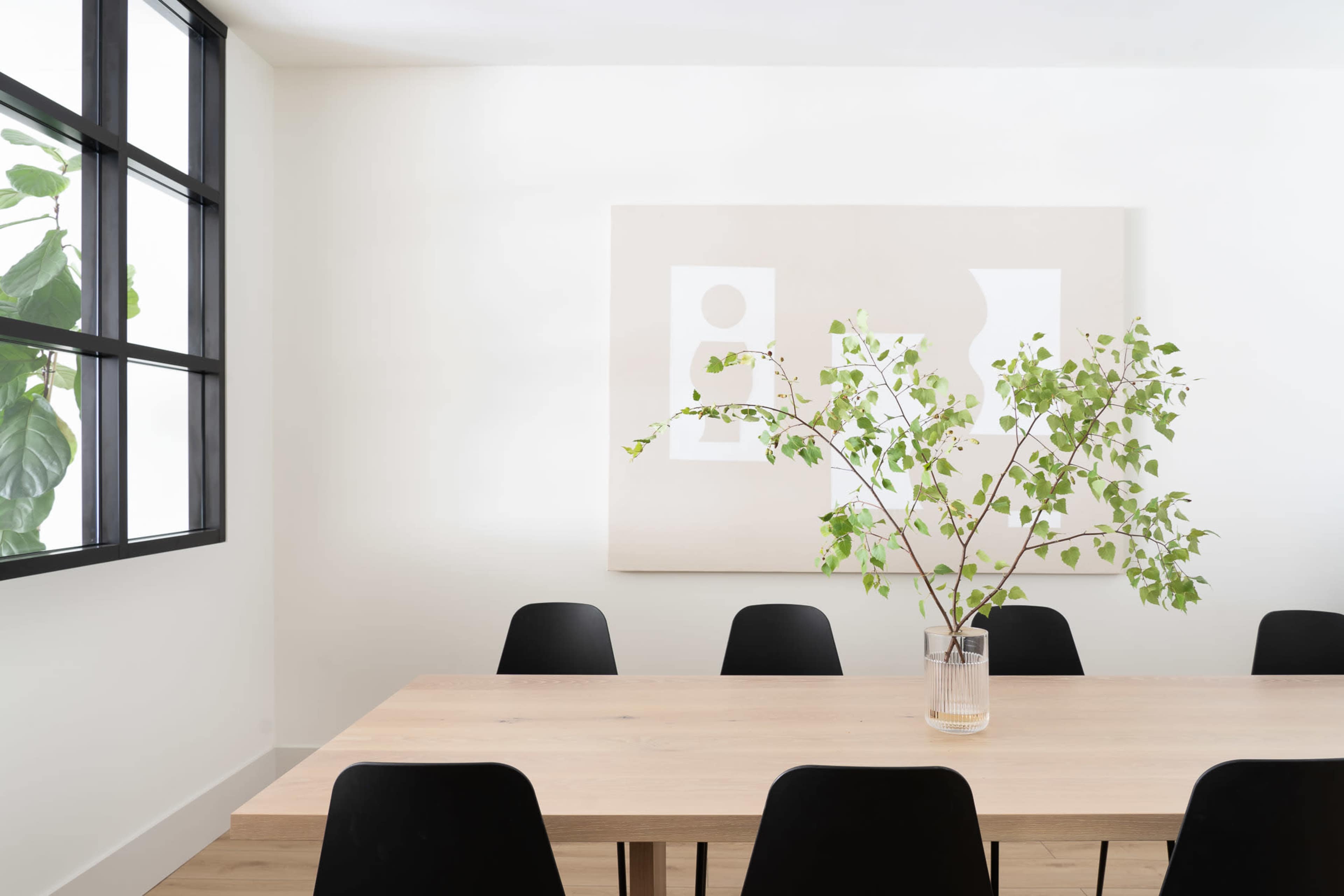 A minimalist dining room features a wooden table surrounded by black chairs and a vase with branches, complemented by a large abstract artwork on the wall.