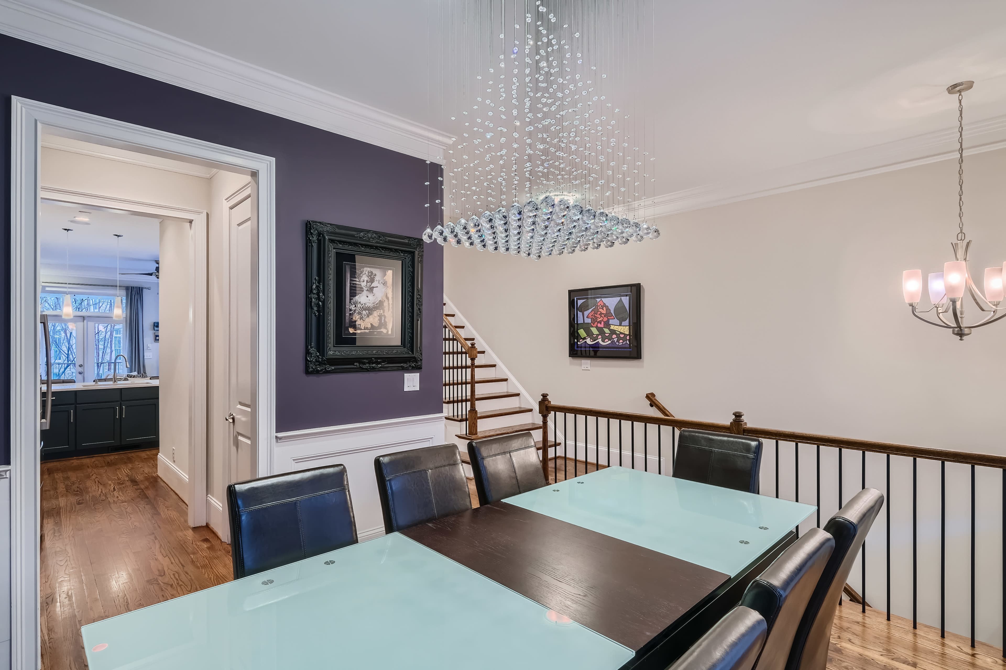 A modern dining room features a long glass table surrounded by black chairs, with a chandelier above and artwork on the walls.