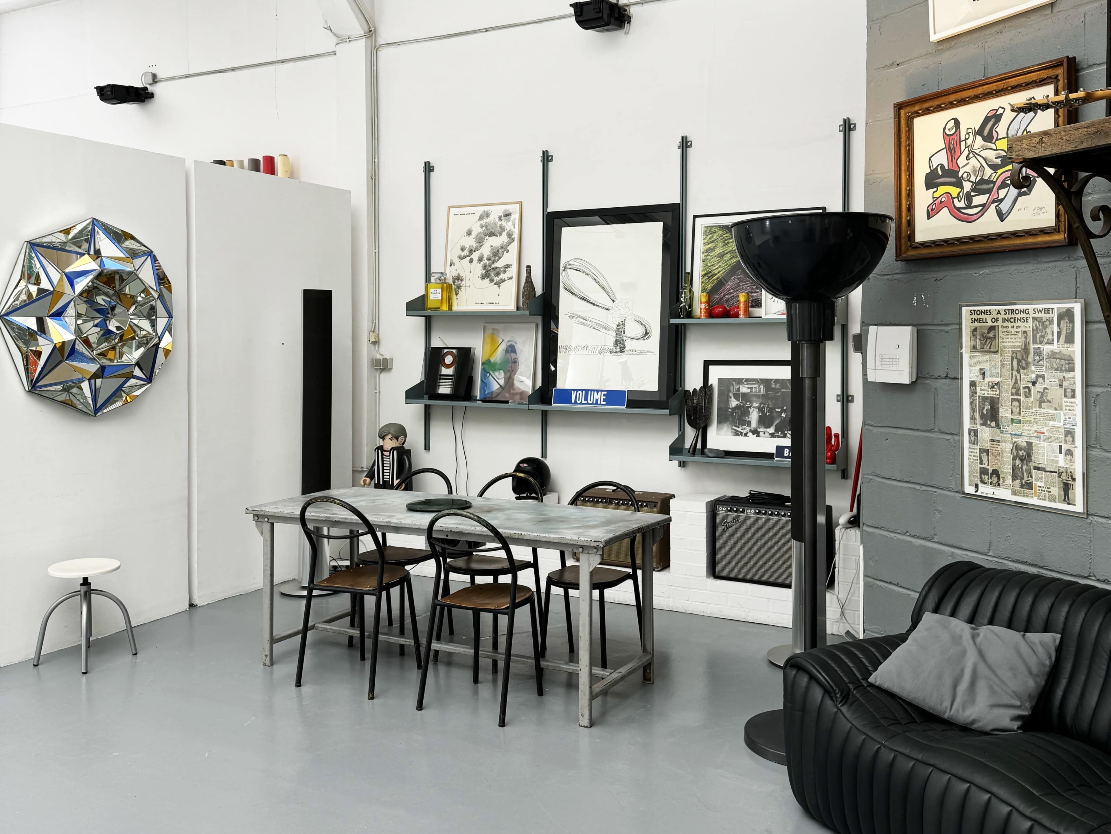 A modern interior workspace with a metal dining table, black chairs, wall-mounted shelves displaying various artworks, and a black leather sofa.