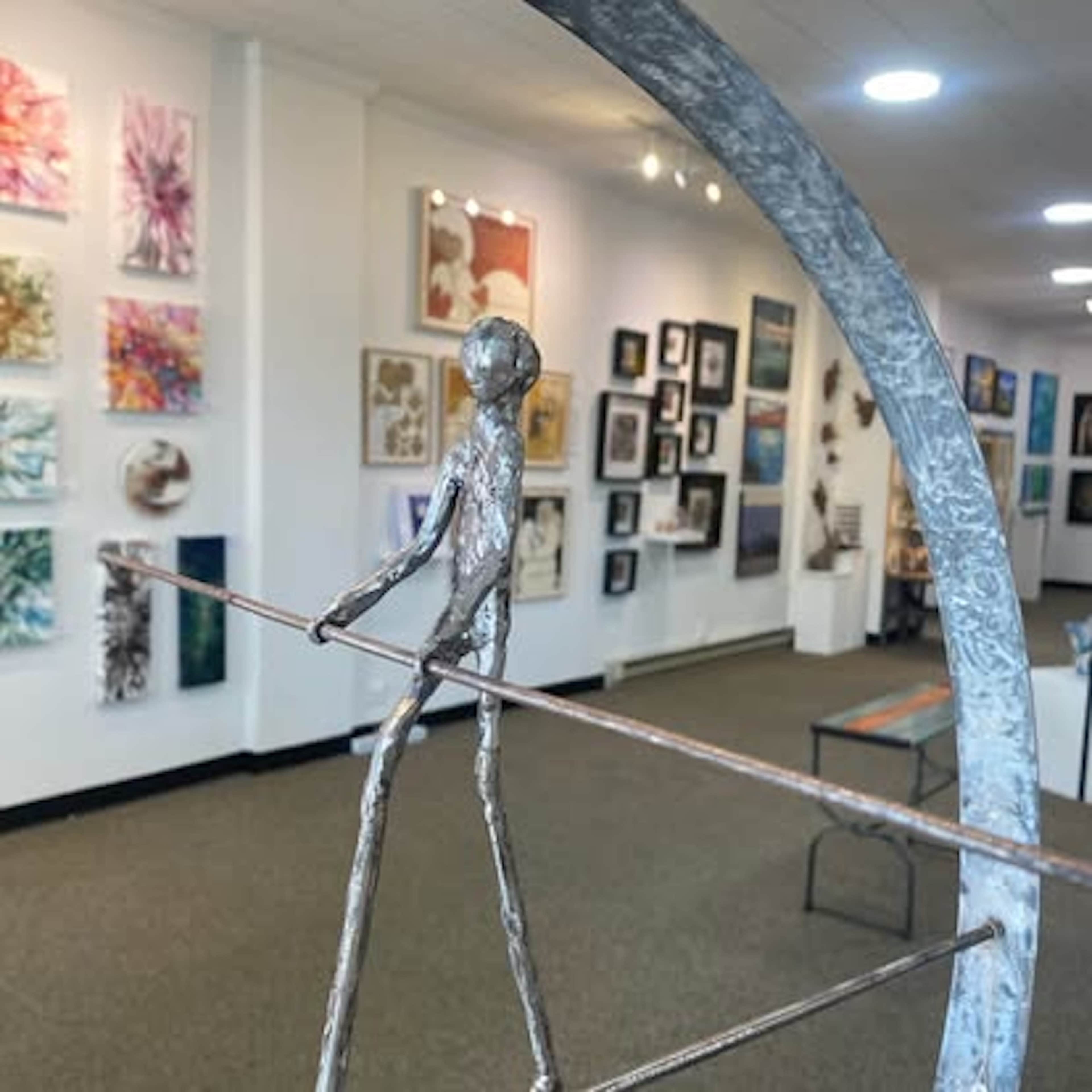 A metal sculpture of a figure balancing on a thin rod is positioned in the foreground of an art gallery featuring various paintings on the walls.