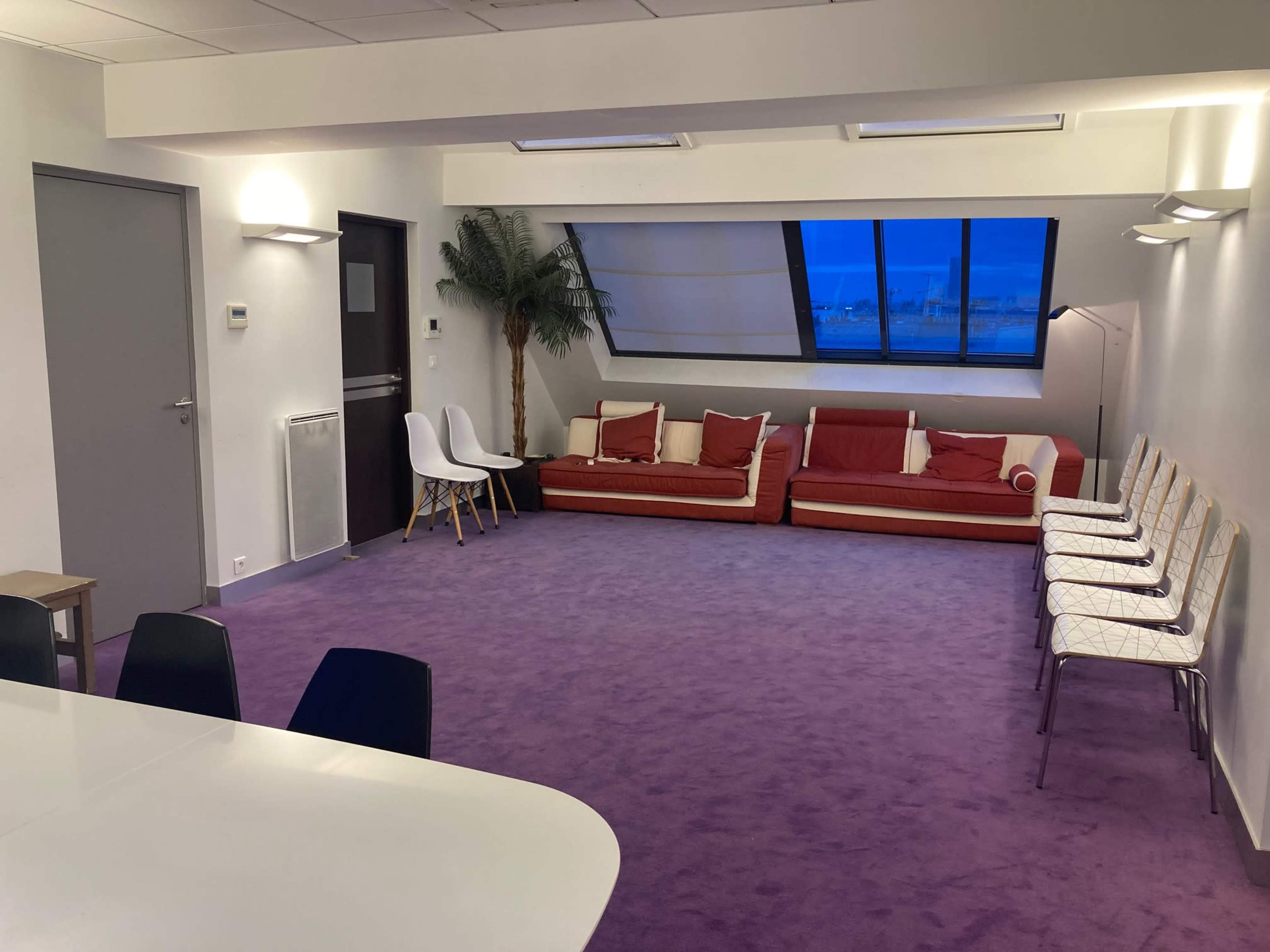 A spacious, modern room with purple carpeting, a large window, a red sofa, and several white chairs arranged along one wall.