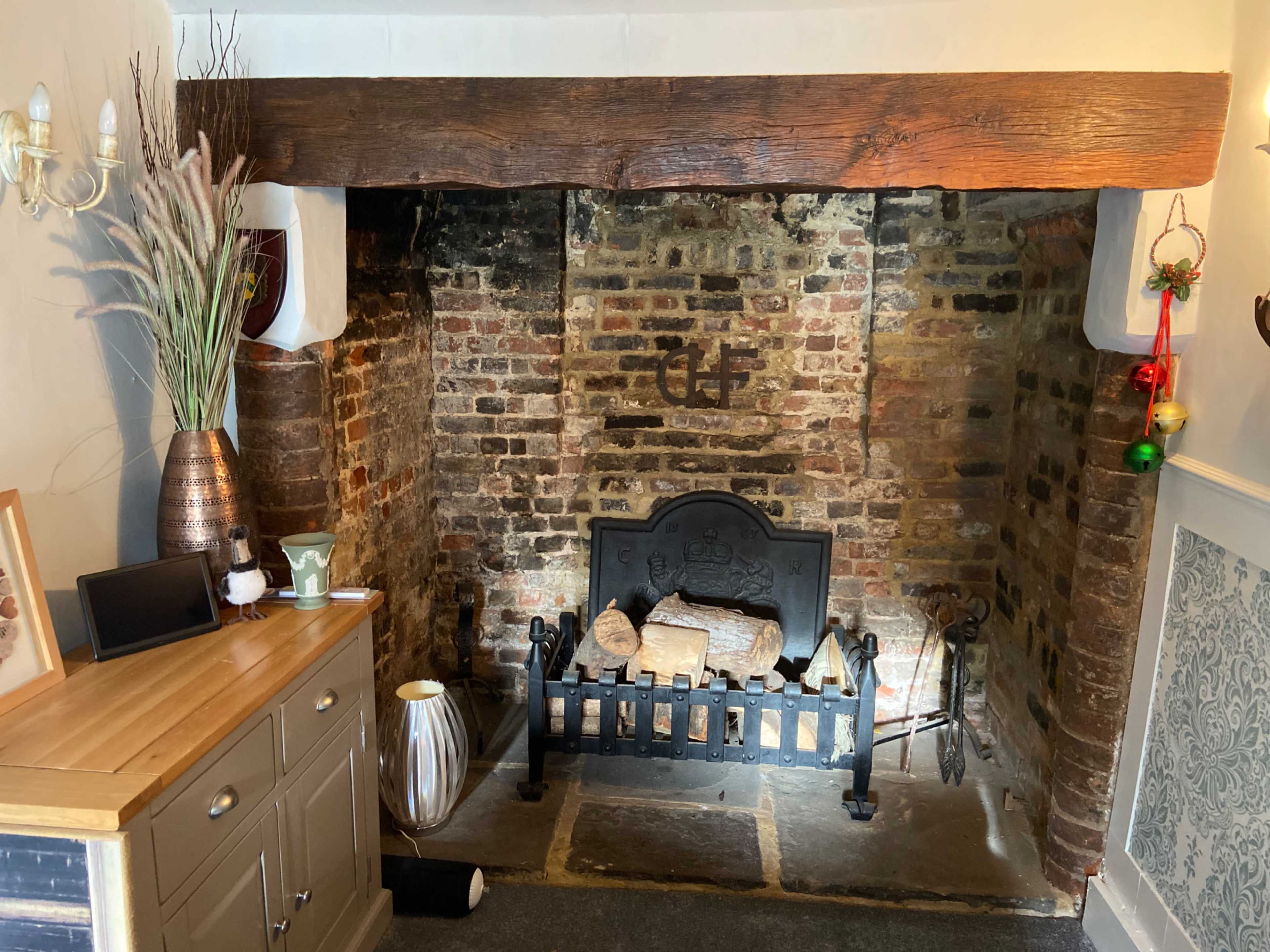 A rustic fireplace with a dark wooden beam above, surrounded by exposed brick walls, and a decorative setup including logs and a metallic vase.